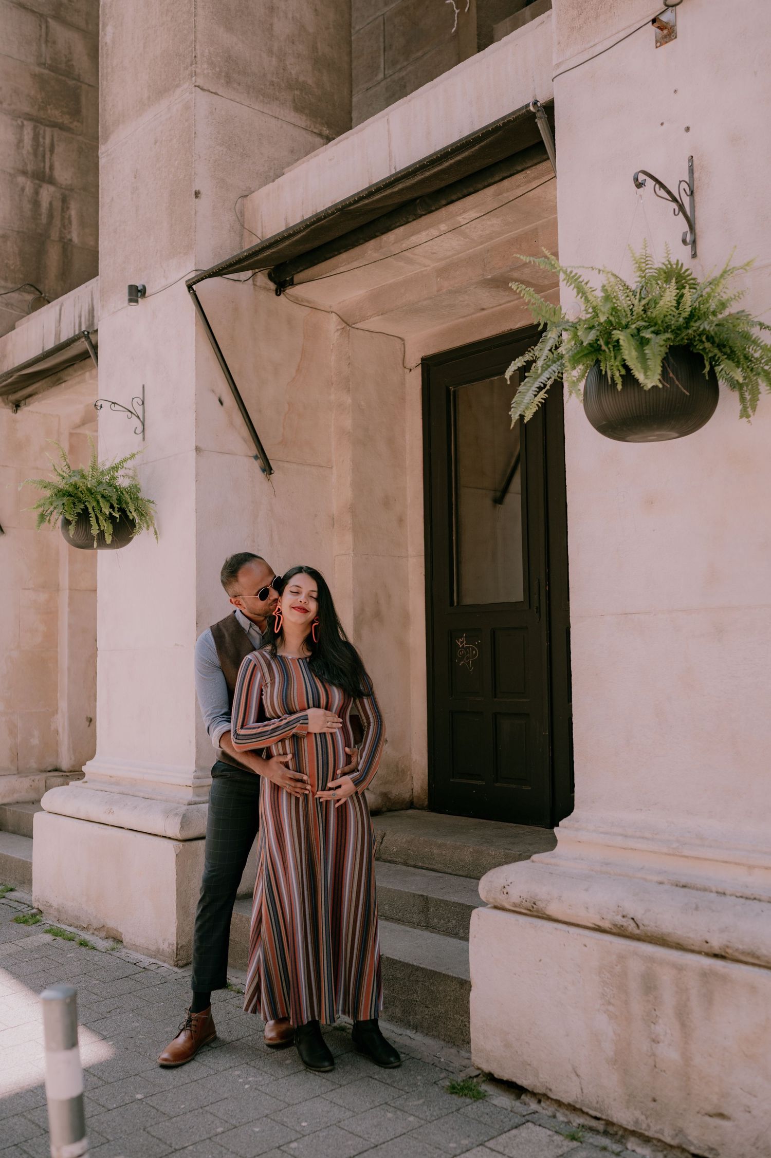 A couple poses in front of a stone building, the woman in a striped maxi dress cradling her baby bump.