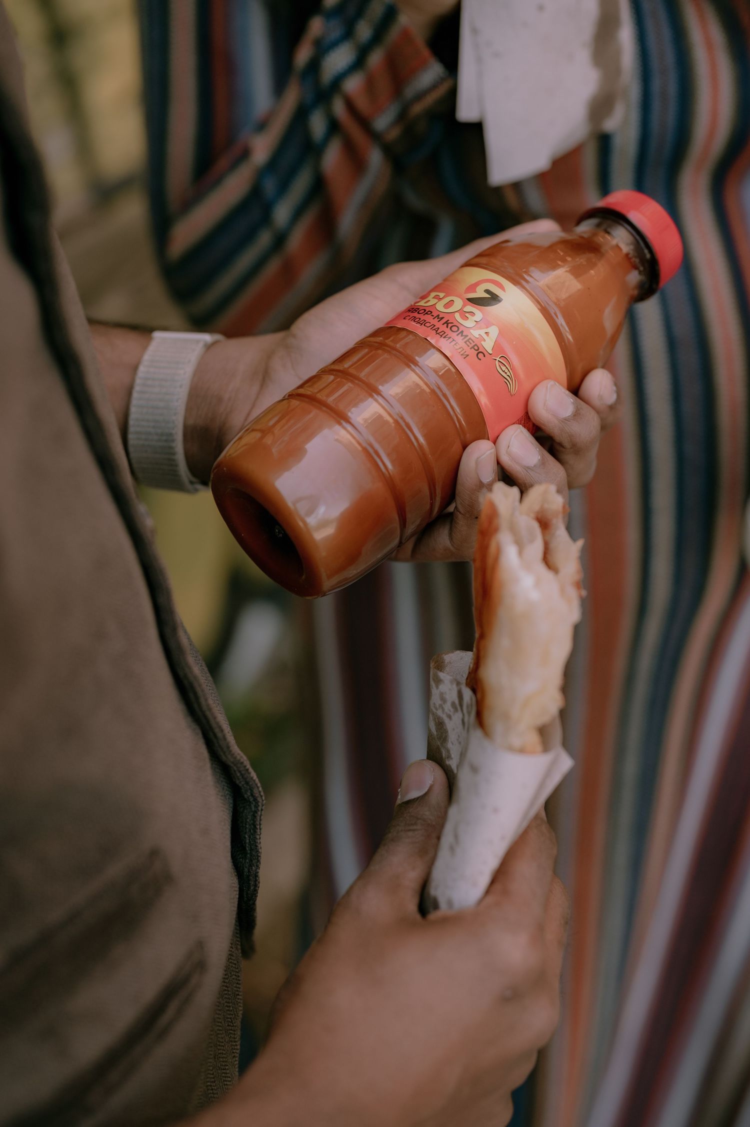 Person pouring orange hot sauce from a bottle onto a food item wrapped in paper outdoors.