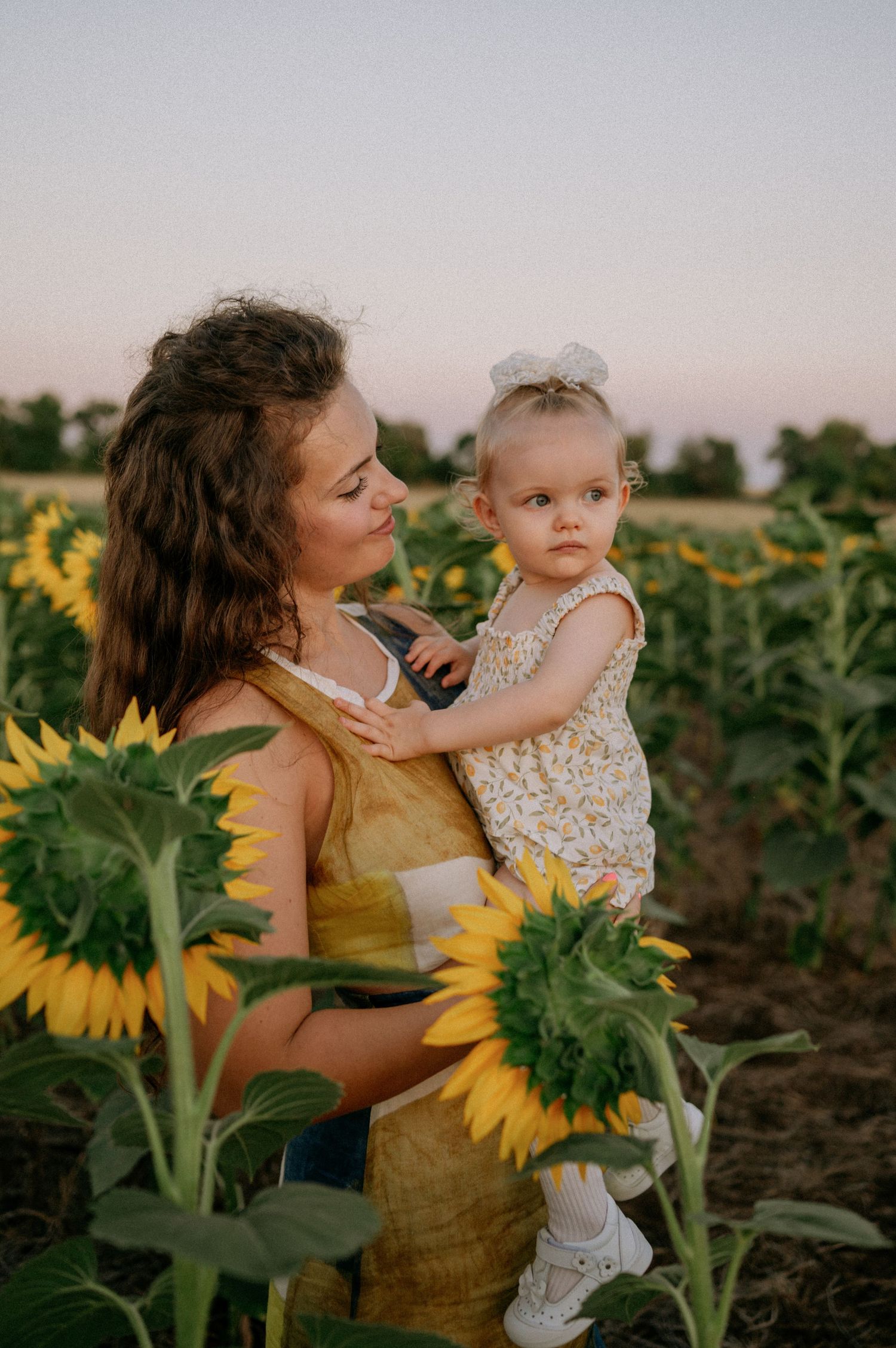 A young mother holds her toddler in a sunflower field at sunset, both wearing yellow outfits.