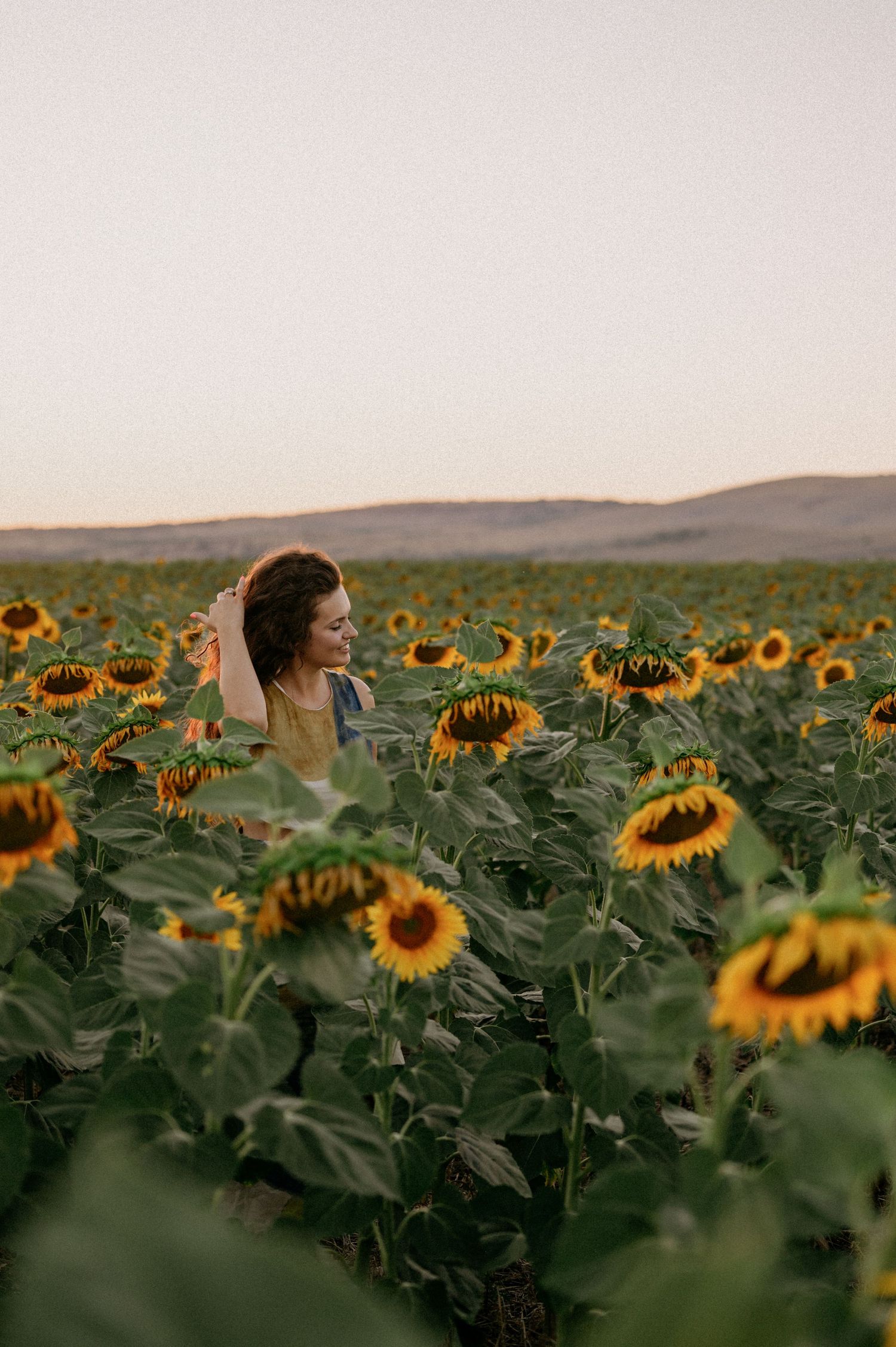 A woman stands smiling in a vast sunflower field at sunset with rolling hills in the background.