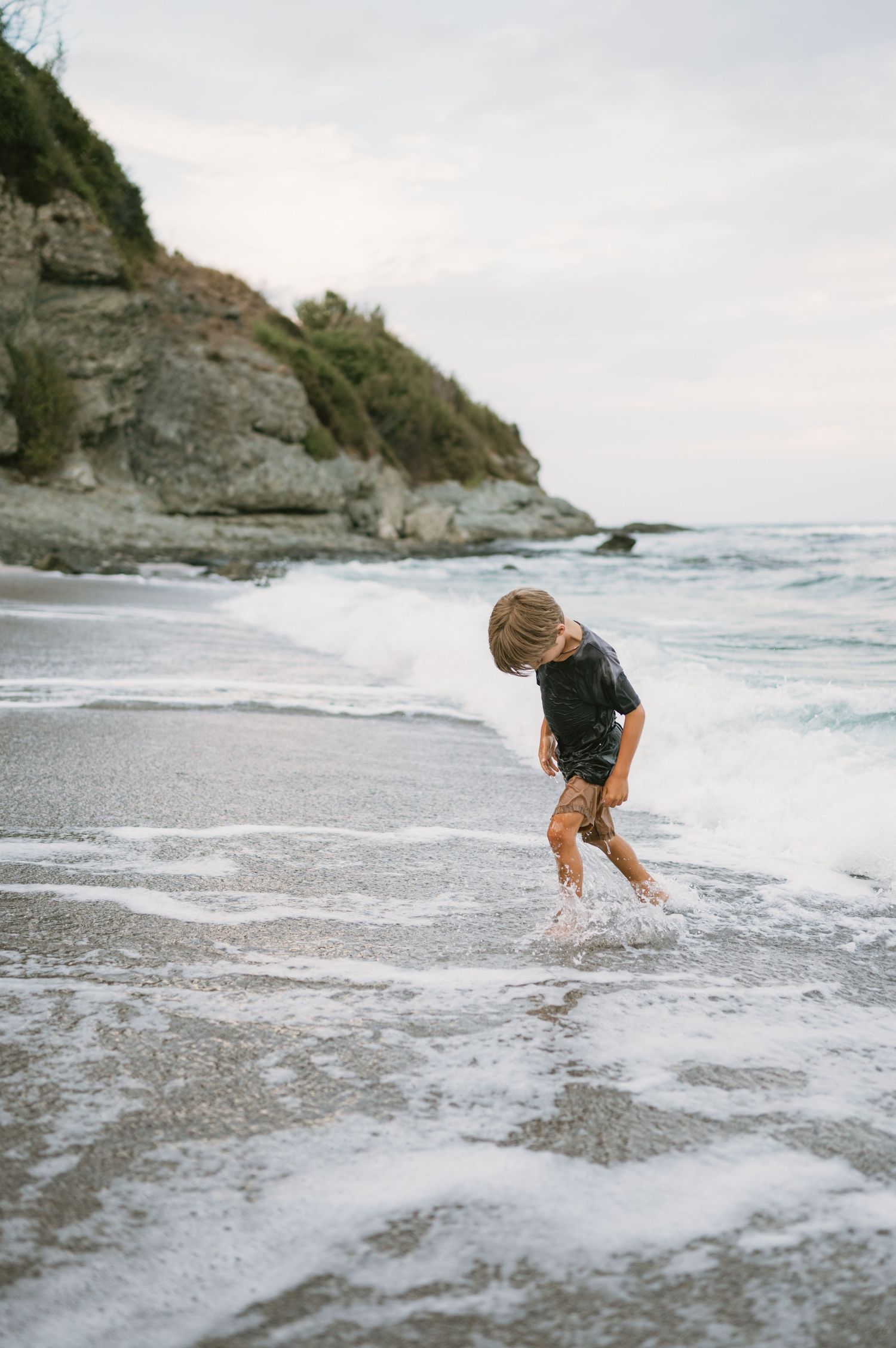 Young boy playing in ocean waves on a rocky beach shore at dusk, wearing dark swimwear.