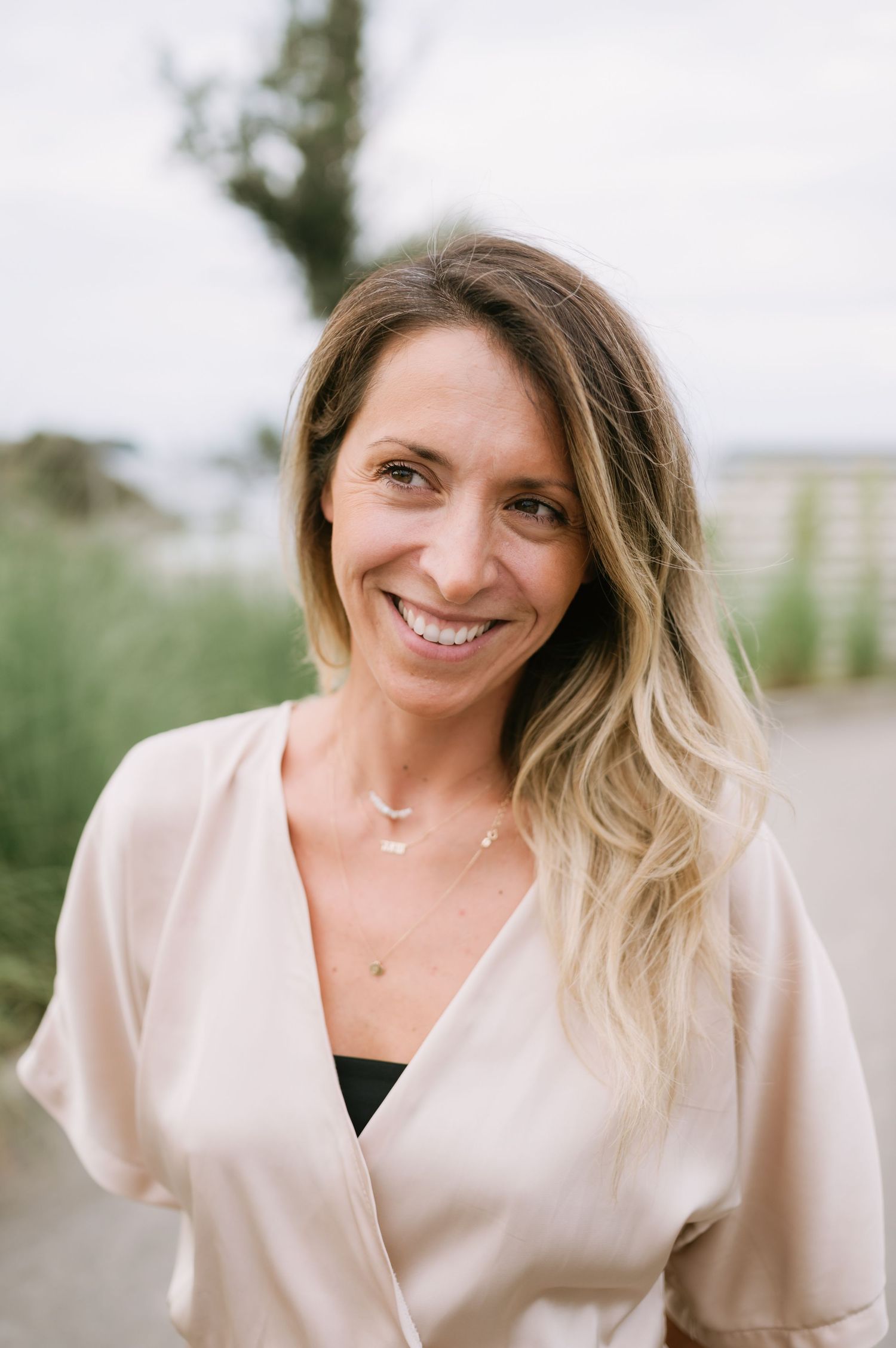 Smiling woman with ombre hair wearing a blush wrap top poses outdoors near greenery.
