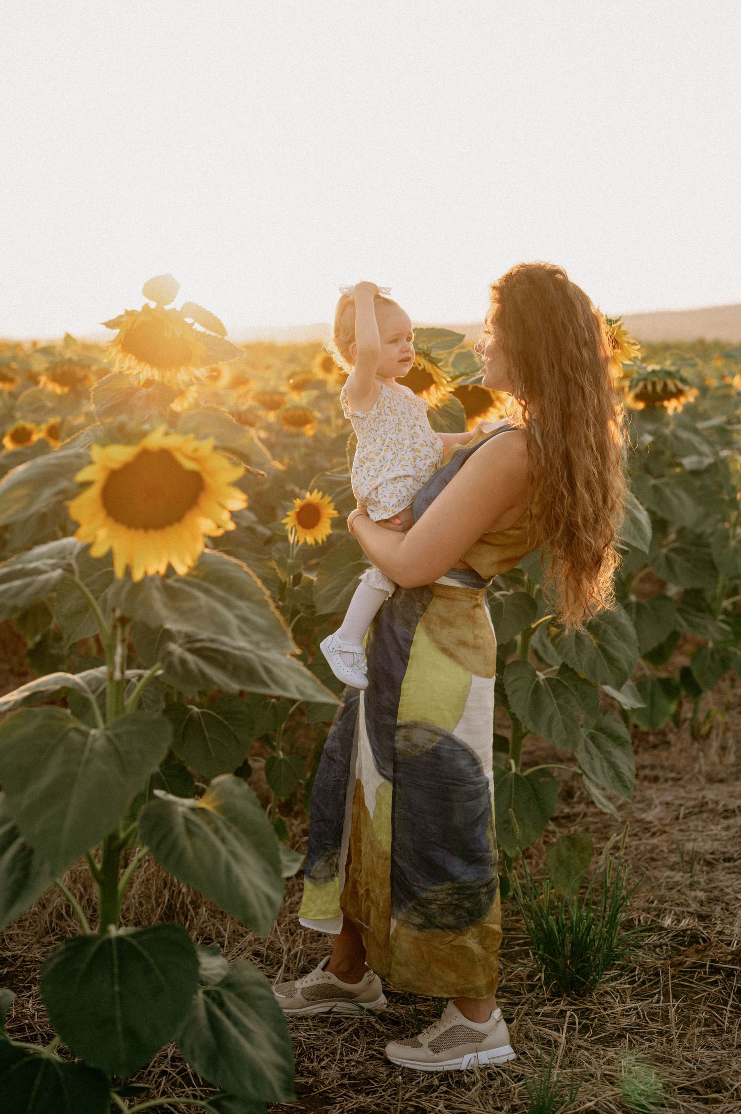 Mother holding baby girl in sunflower field at golden hour, wearing colorful maxi dress.