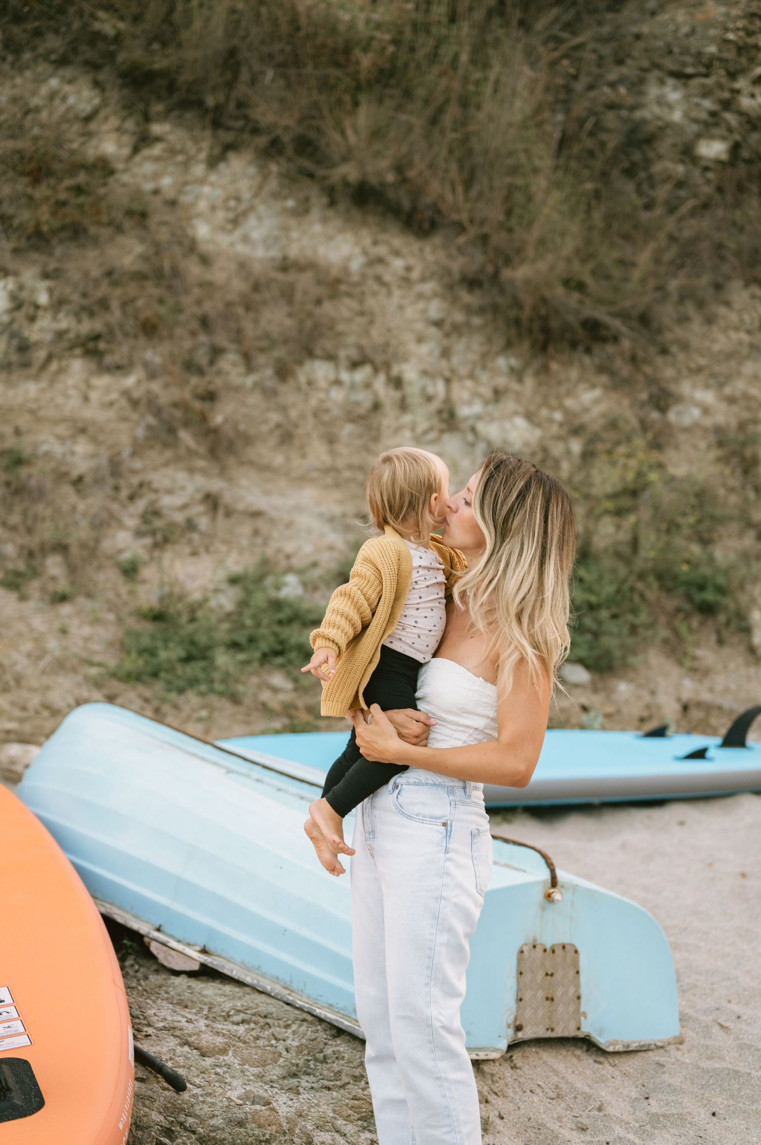 A mother holds and kisses her toddler on a sandy beach with colorful kayaks and a grassy hillside backdrop.