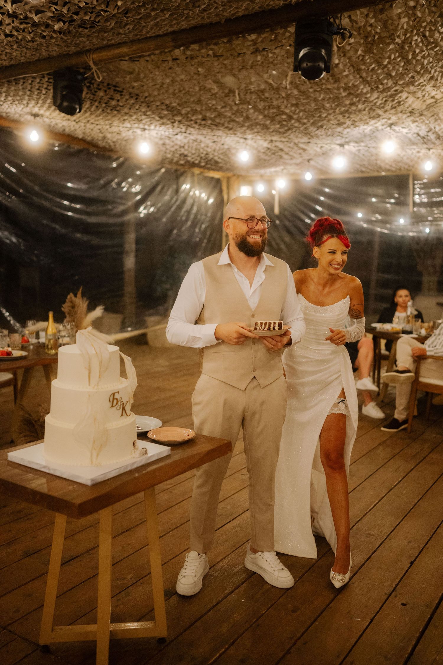 A wedding reception on a rustic wooden deck with string lights overhead and a white wedding cake display.