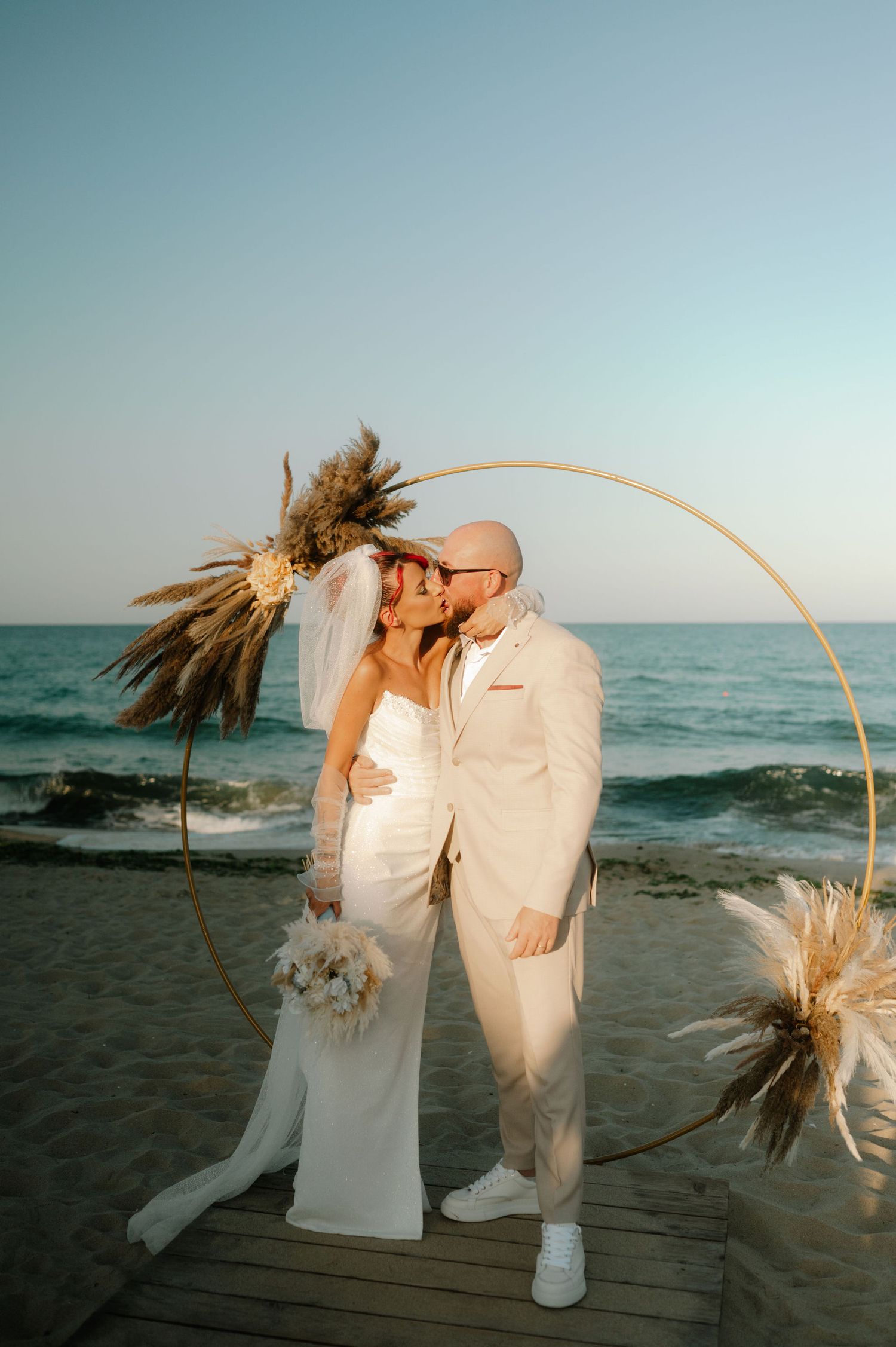 A couple shares a romantic kiss on the beach at sunset with decorative dried pampas grass arrangements framing the scene.