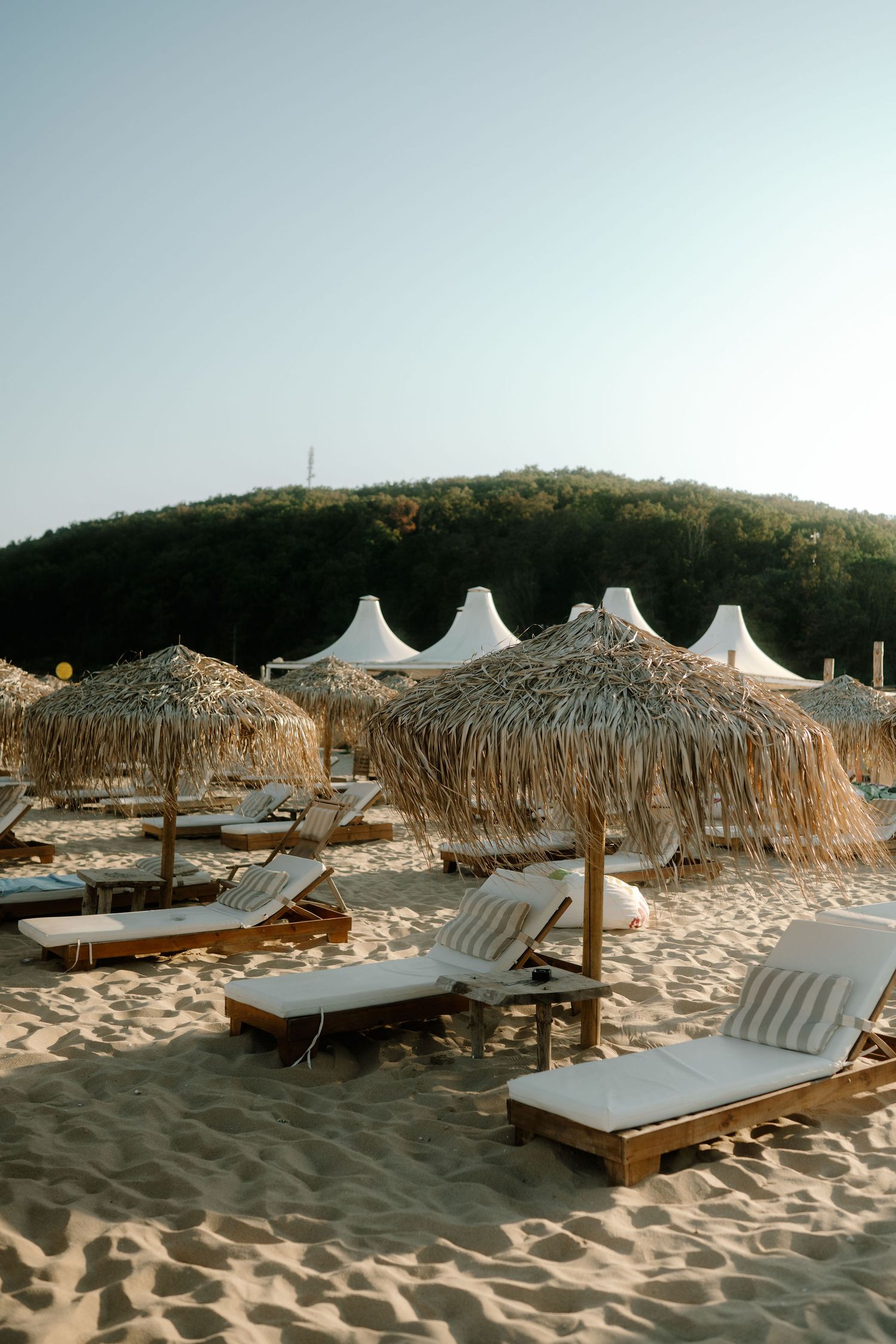 Thatched beach umbrellas and white lounge chairs line a sandy beach with white tents and hills in the background.