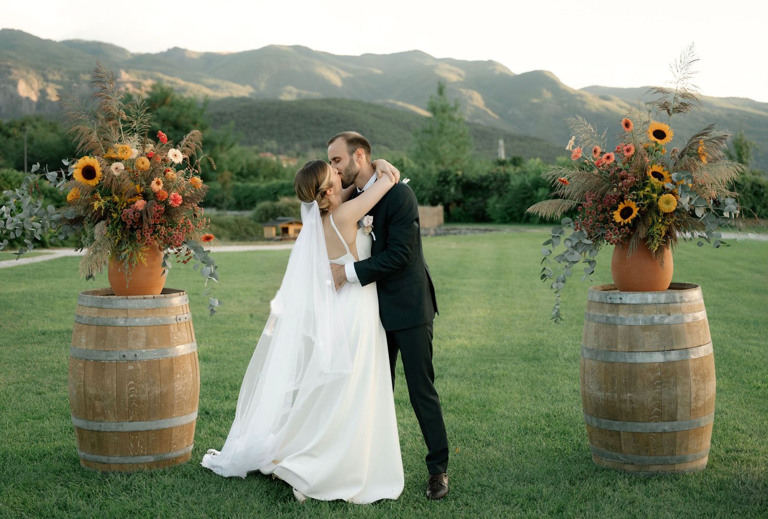 Romantic couple embraces near rustic wine barrels in a scenic mountain vineyard setting at sunset.