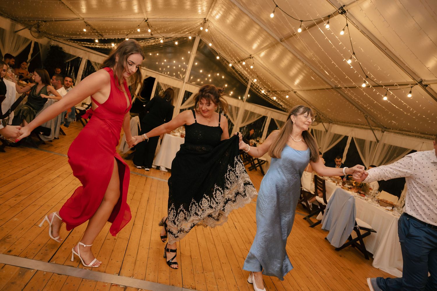 Wedding guests dance and celebrate on a rustic wooden dance floor under string lights at an indoor reception venue.