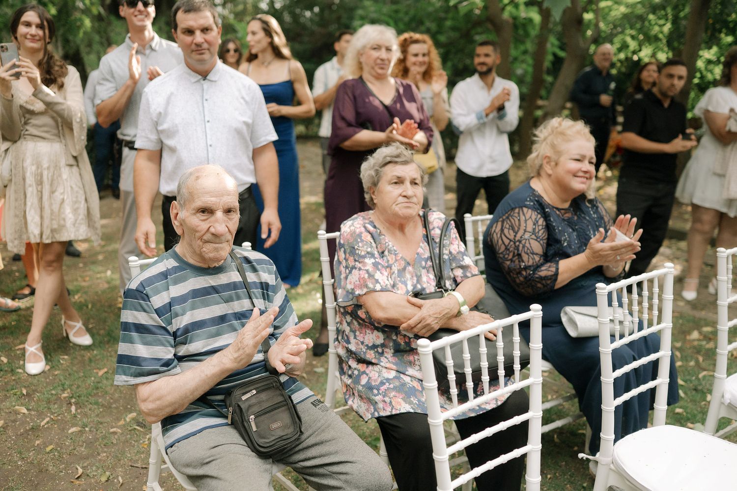Wedding guests seated in white chairs at an outdoor garden ceremony applaud during a special moment.