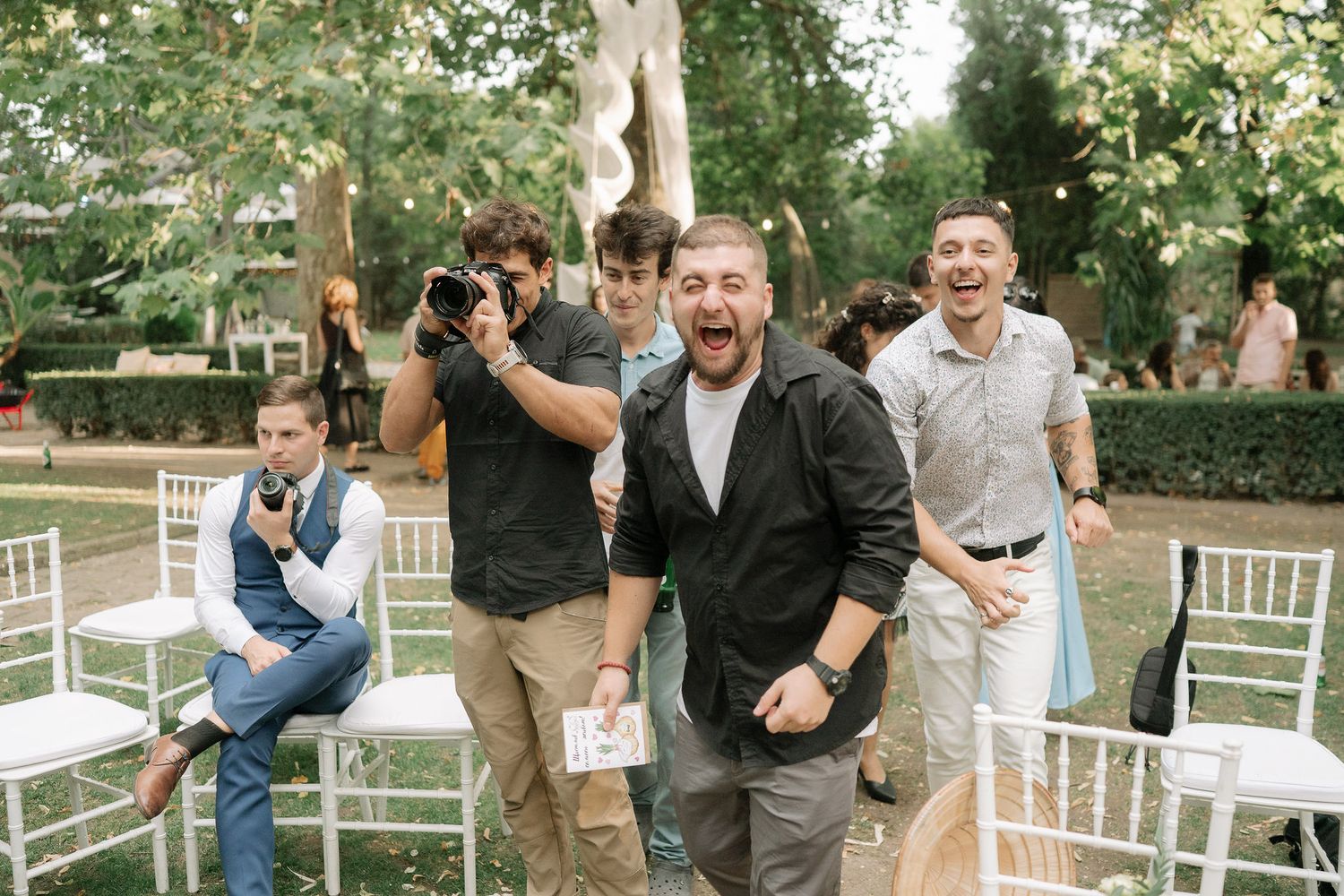 A group celebrates joyfully outdoors at a wedding venue with white chairs and natural greenery in the background.