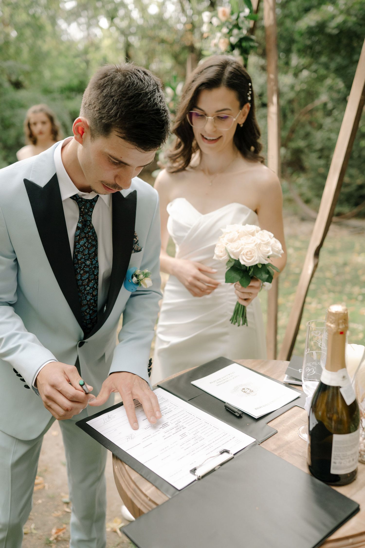Couple signing marriage documents at outdoor wedding ceremony with floral bouquet and grey suit details.