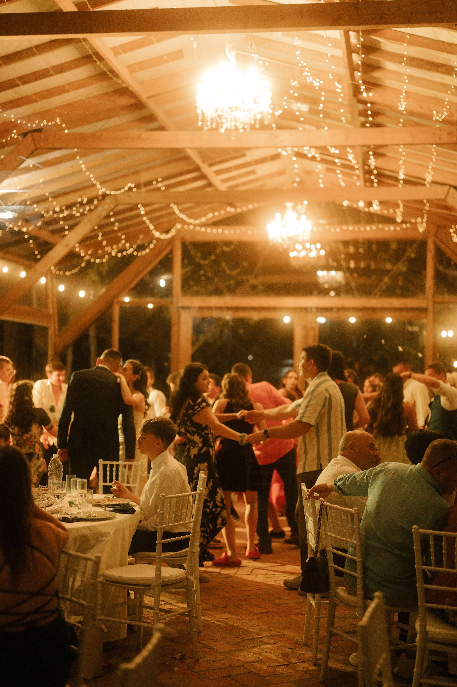 Wedding reception guests dancing and dining under a rustic pavilion with twinkling lights and chandeliers at night.