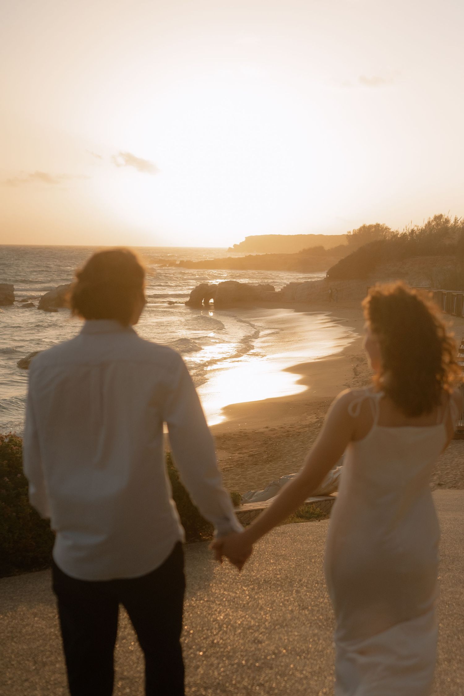 Couple holding hands watching a romantic golden sunset over a rocky beach shoreline.