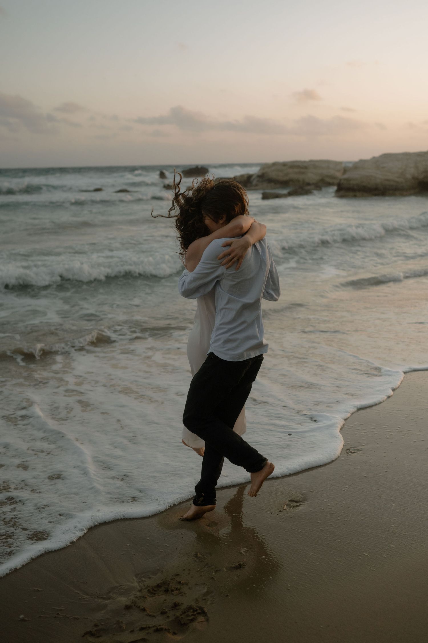 Two people hugging barefoot on a wet sandy beach shore at dusk with rocky coastline.