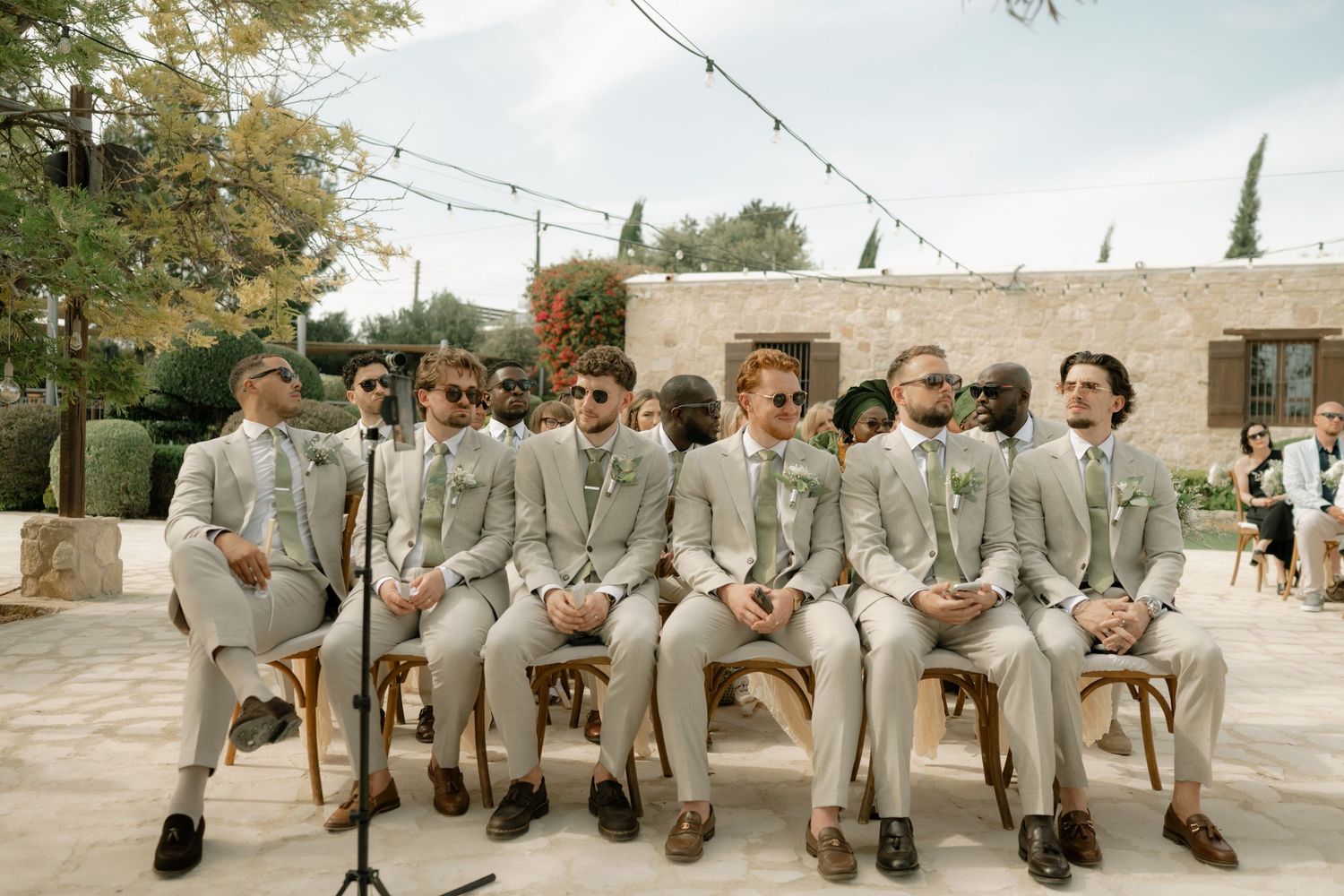 Groomsmen in matching gray suits seated outdoors at a wedding ceremony with string lights overhead.