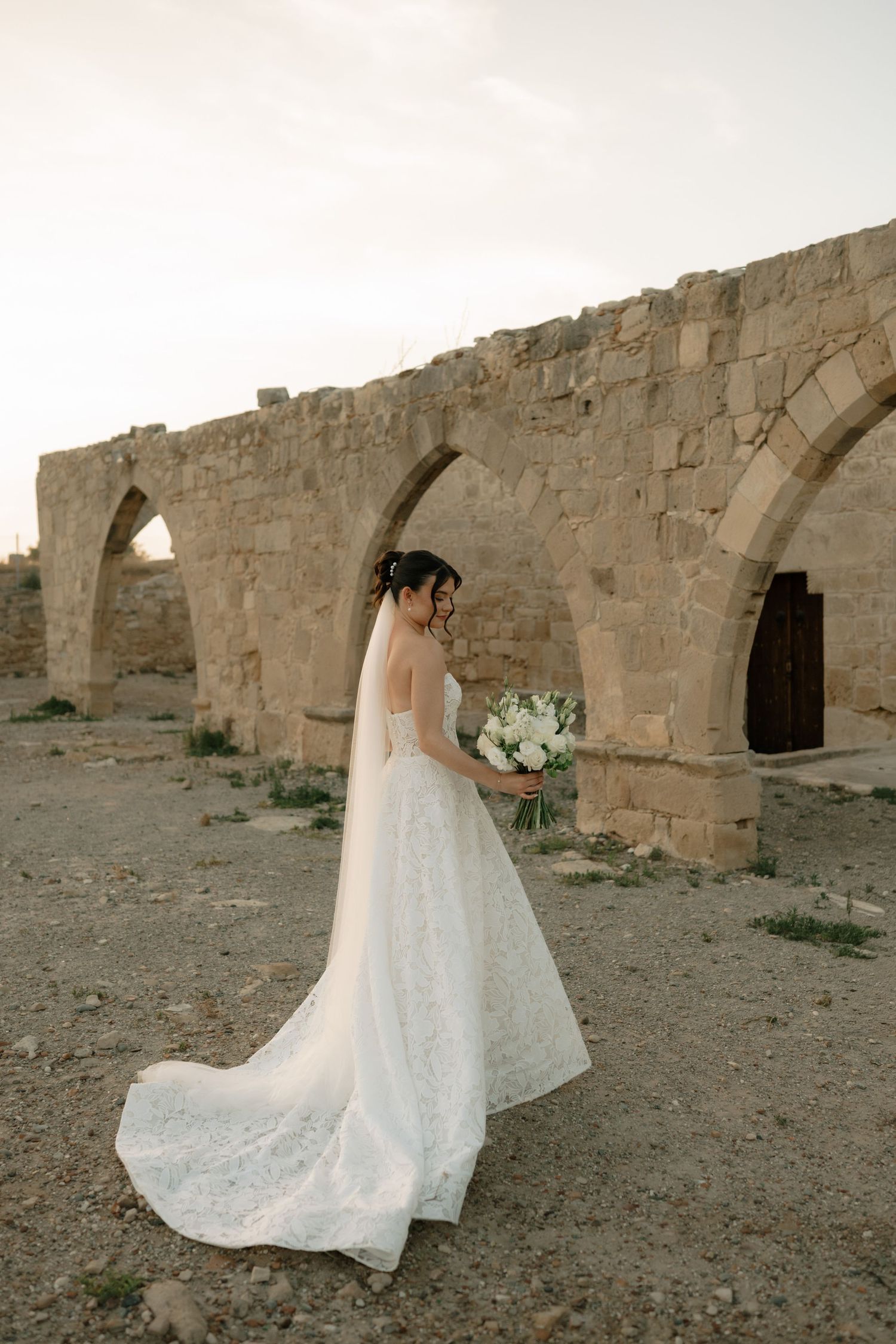 Bride in white lace gown with veil holds bouquet before ancient stone arches at sunset.