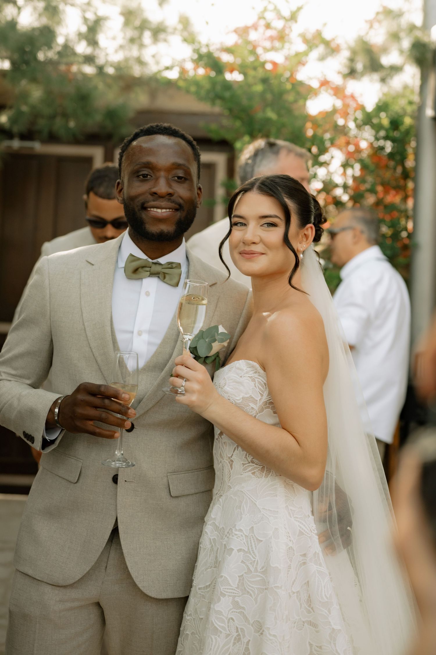 Smiling newlyweds in elegant wedding attire holding champagne flutes at an outdoor wedding reception.