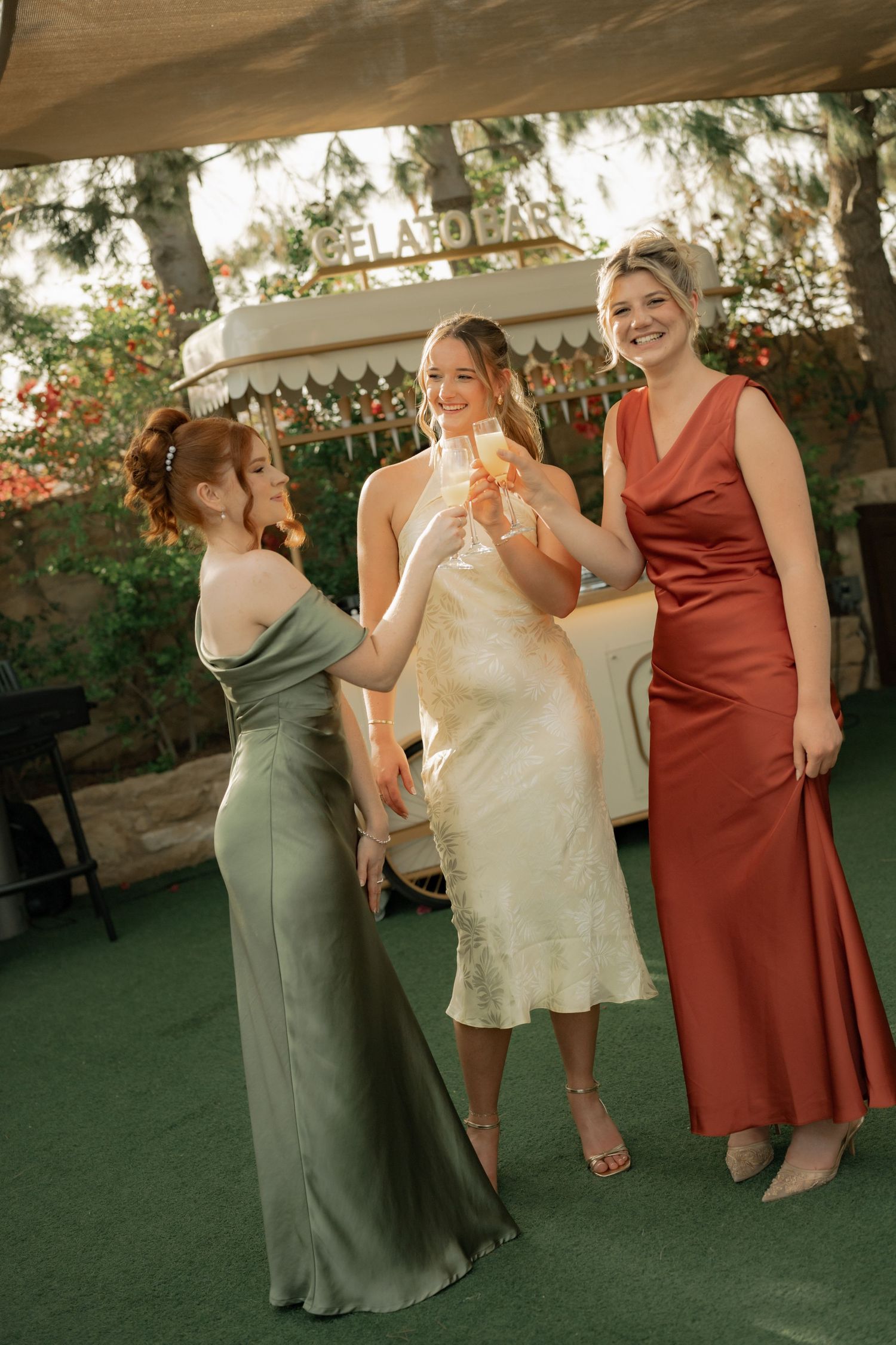 Three women in elegant gowns toast with champagne glasses at an outdoor wedding celebration near a floral bar.