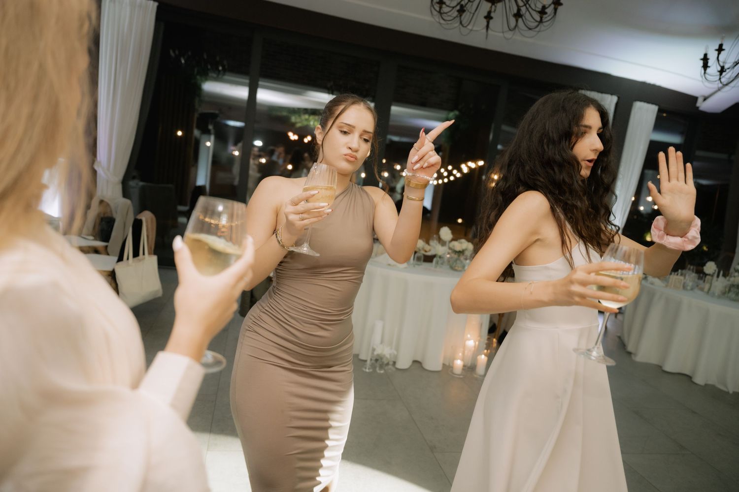 A person in a brown dress and another in a white dress dance together in an indoor venue with chandeliers.