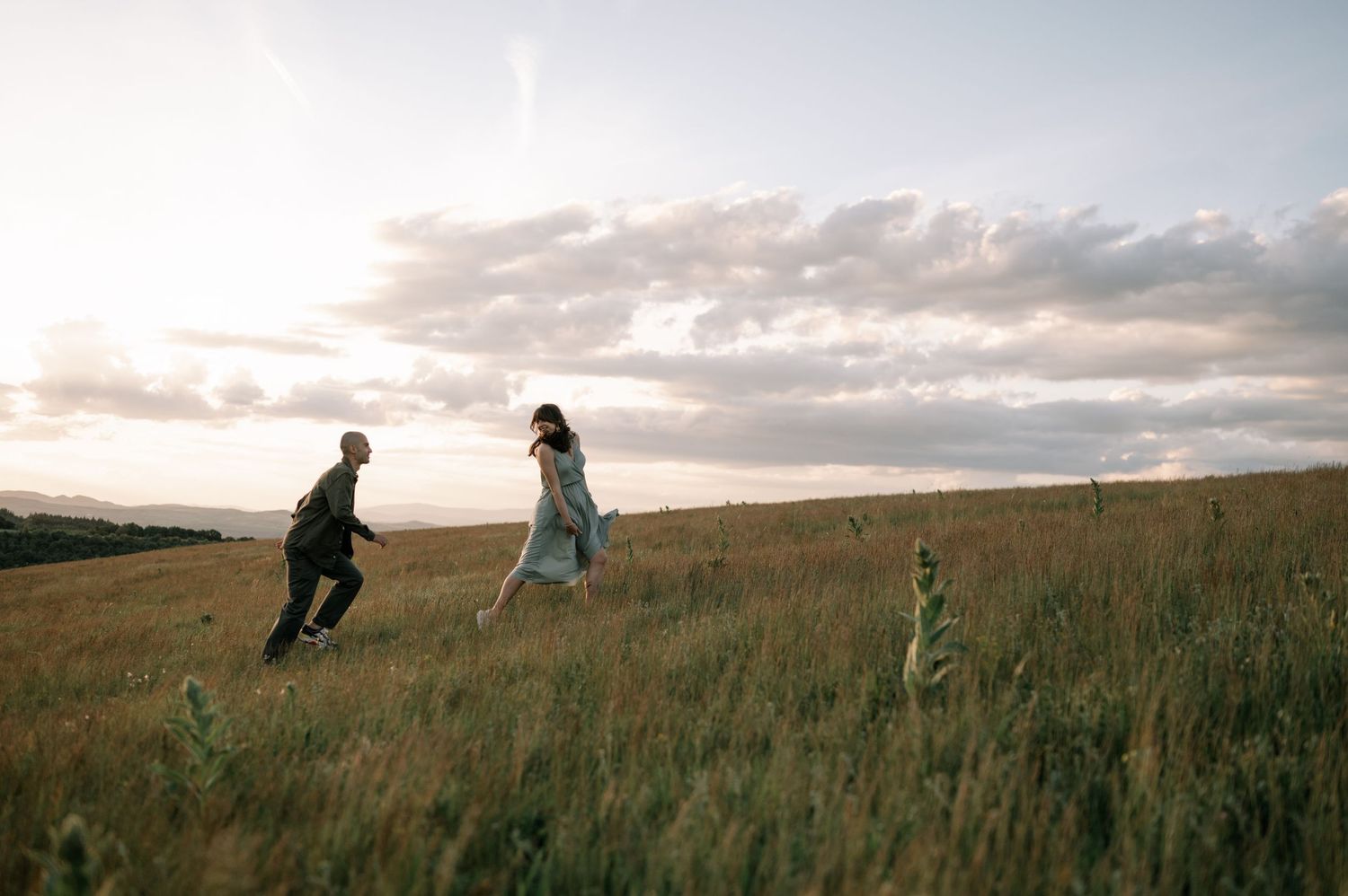 Couple running through golden field at sunset under dramatic cloudy sky in romantic outdoor photography.