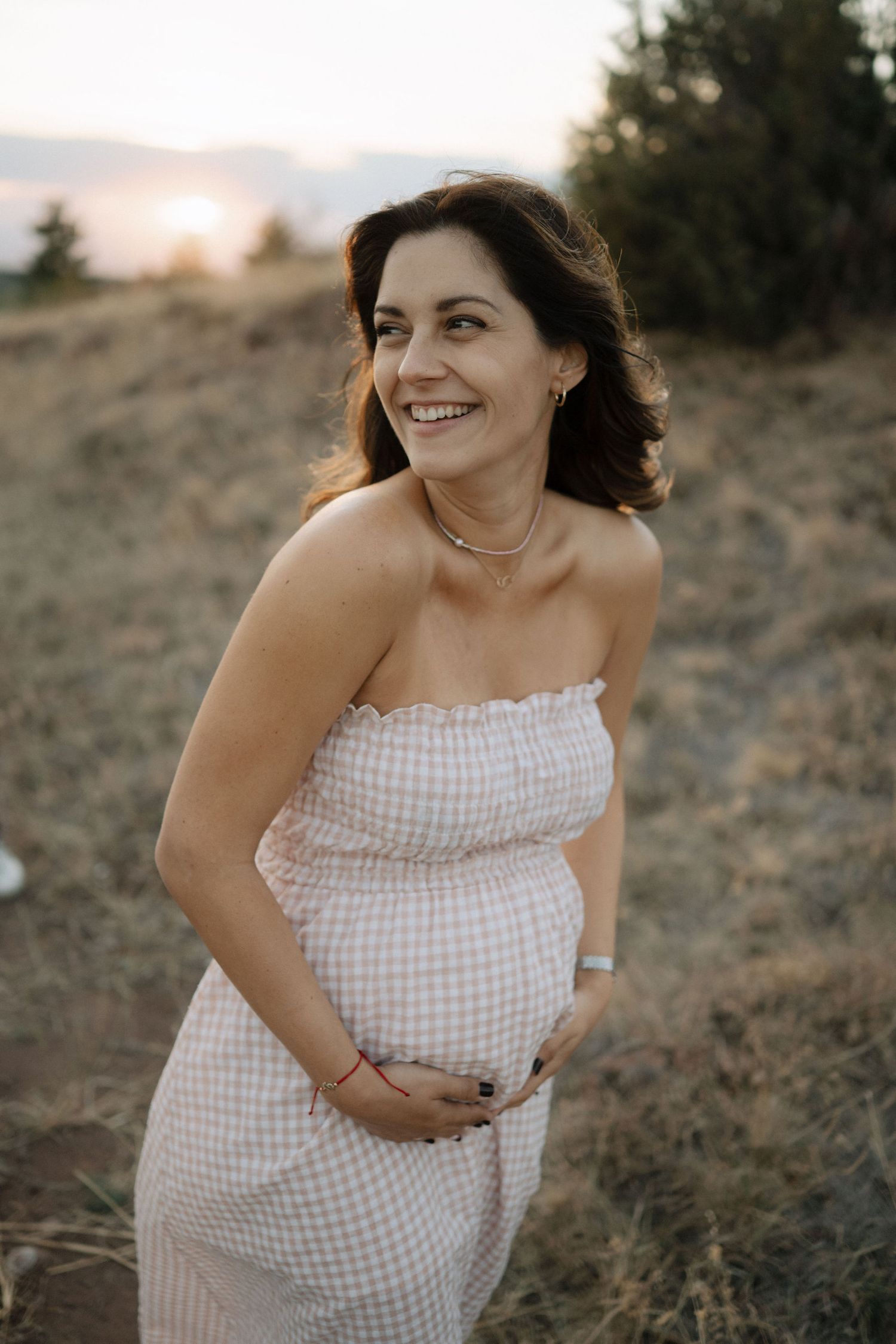 A person in a white gingham dress poses outdoors during golden hour on a grassy hillside.