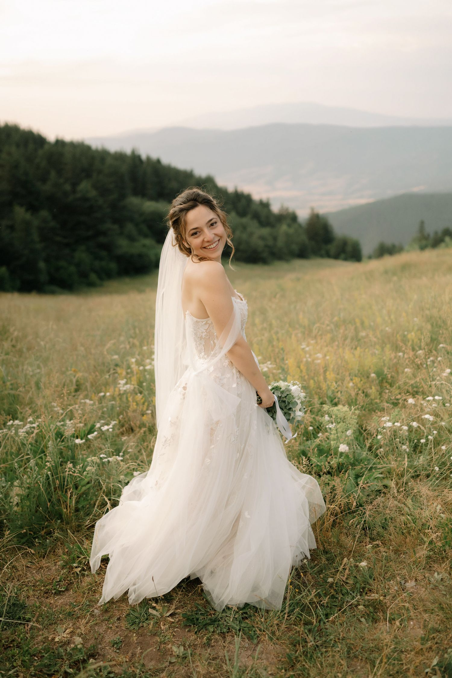 A bride in a flowing white dress twirls gracefully in a wildflower meadow with misty mountains in the background.