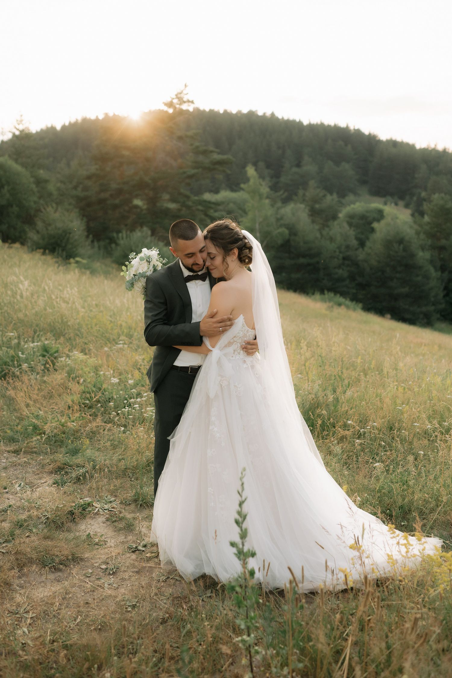 A romantic sunset wedding photo series shows a couple embracing in a golden meadow with evergreen forest in the background.