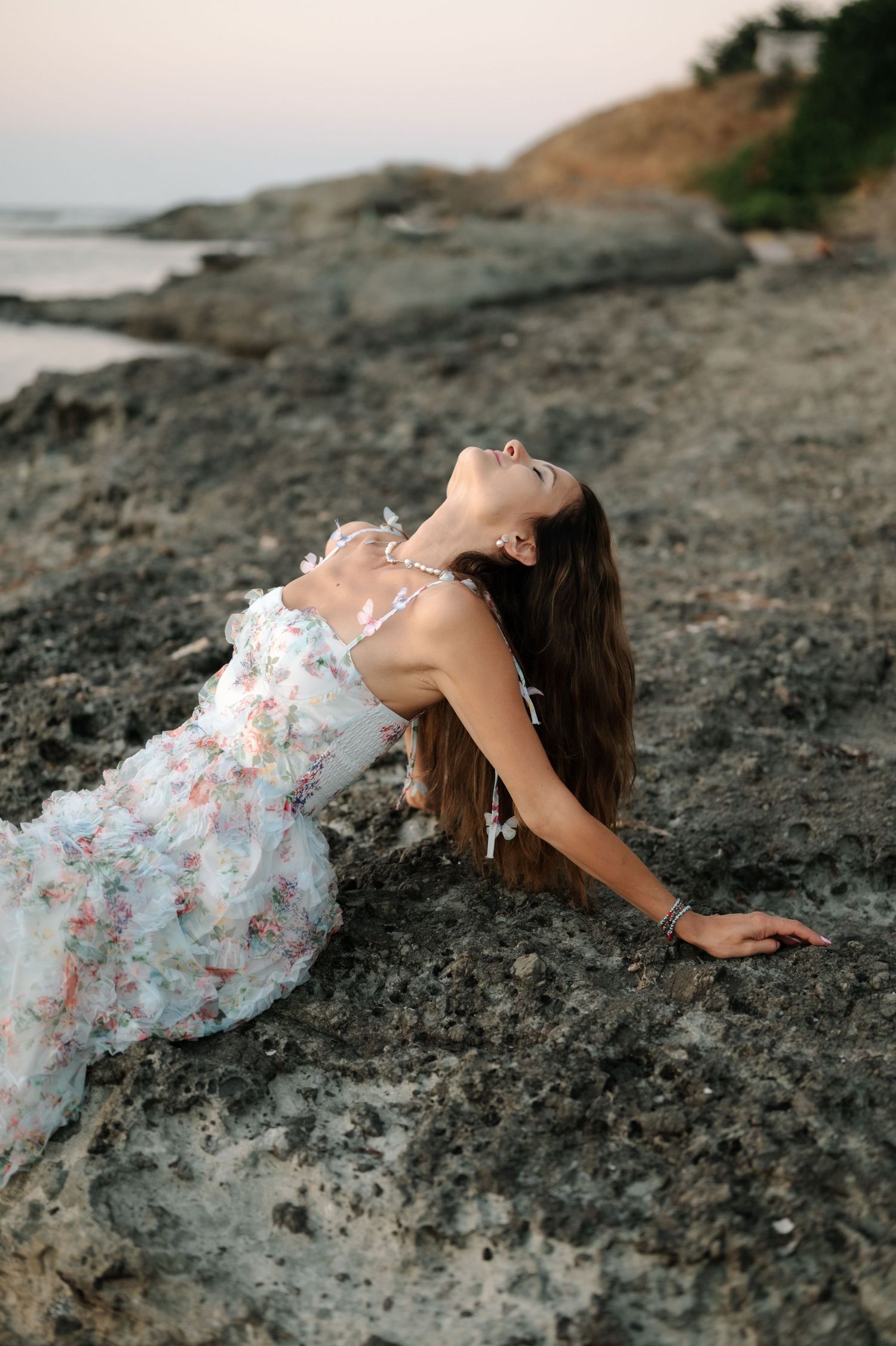 A person in a white floral dress lies gracefully on dark rocky terrain by the coastline.