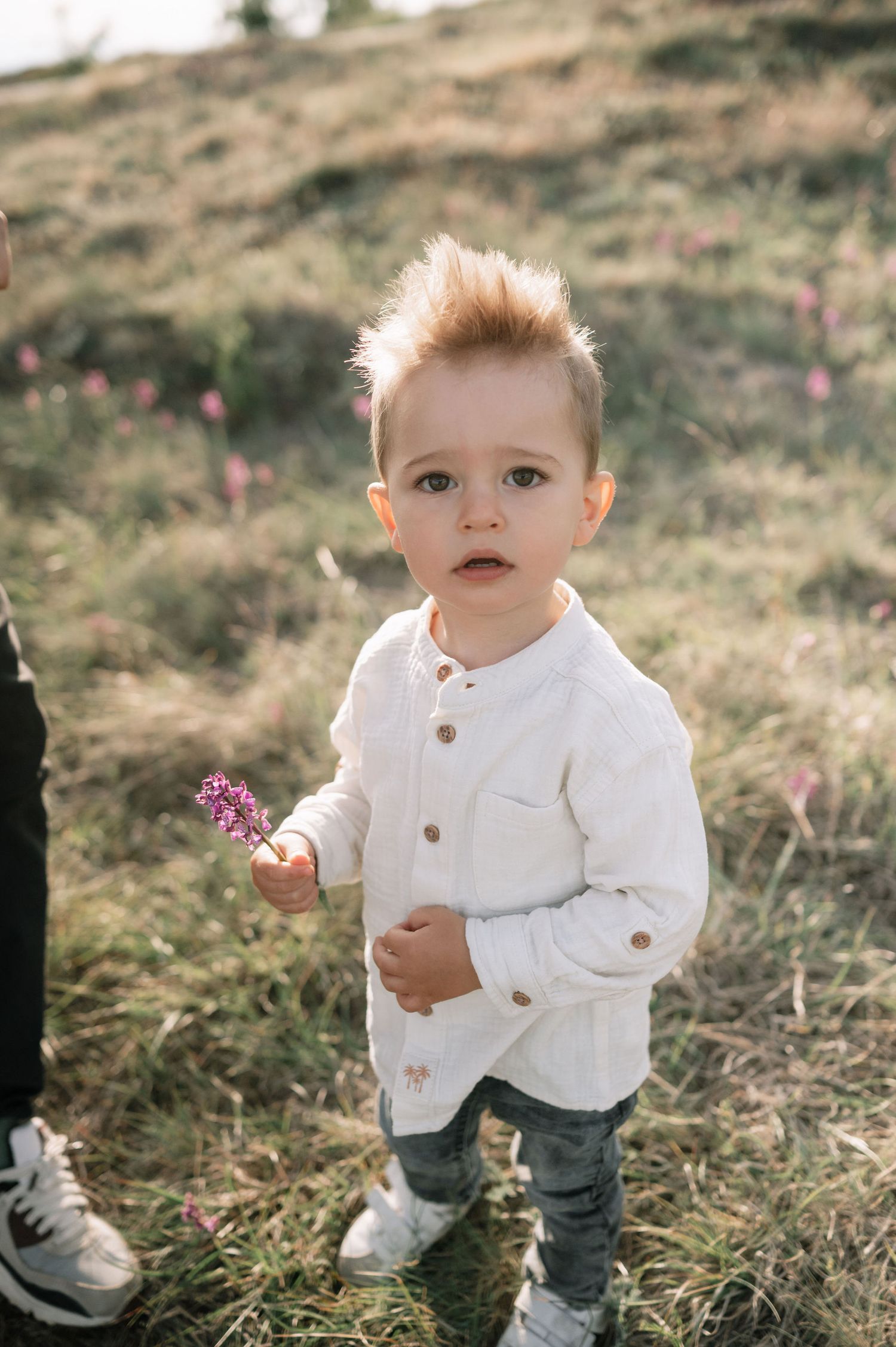 A young child in a white sweater and jeans explores a grassy hillside on a sunny day.