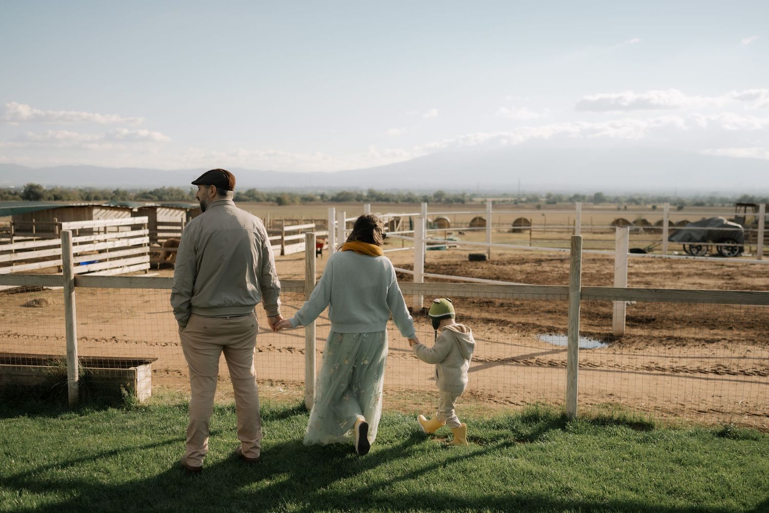 Three people stand at a wooden fence overlooking a rural farm landscape with mountains in the distance.