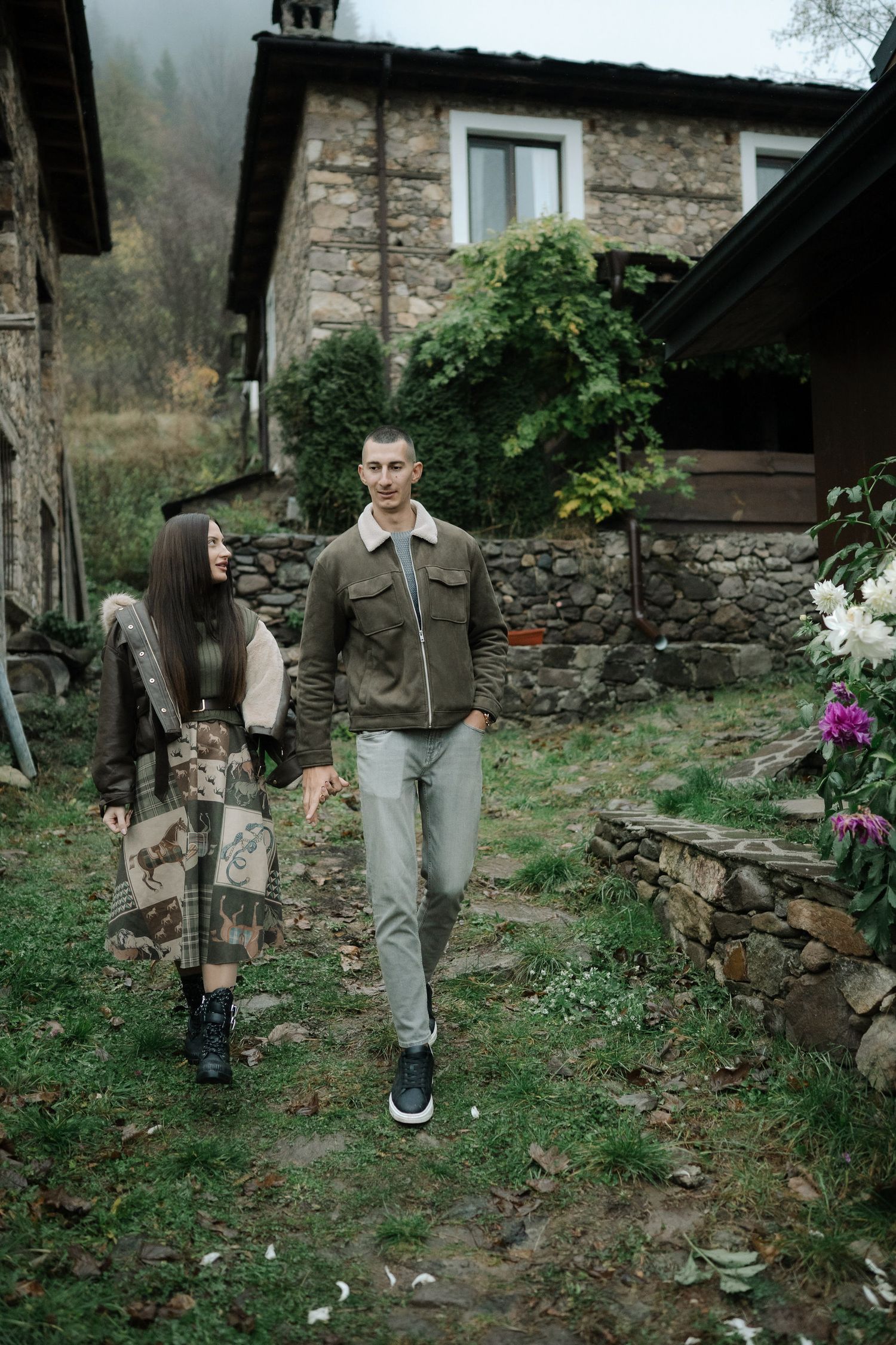 Person walking along a stone wall path in a rustic garden setting with scattered debris and vegetation.