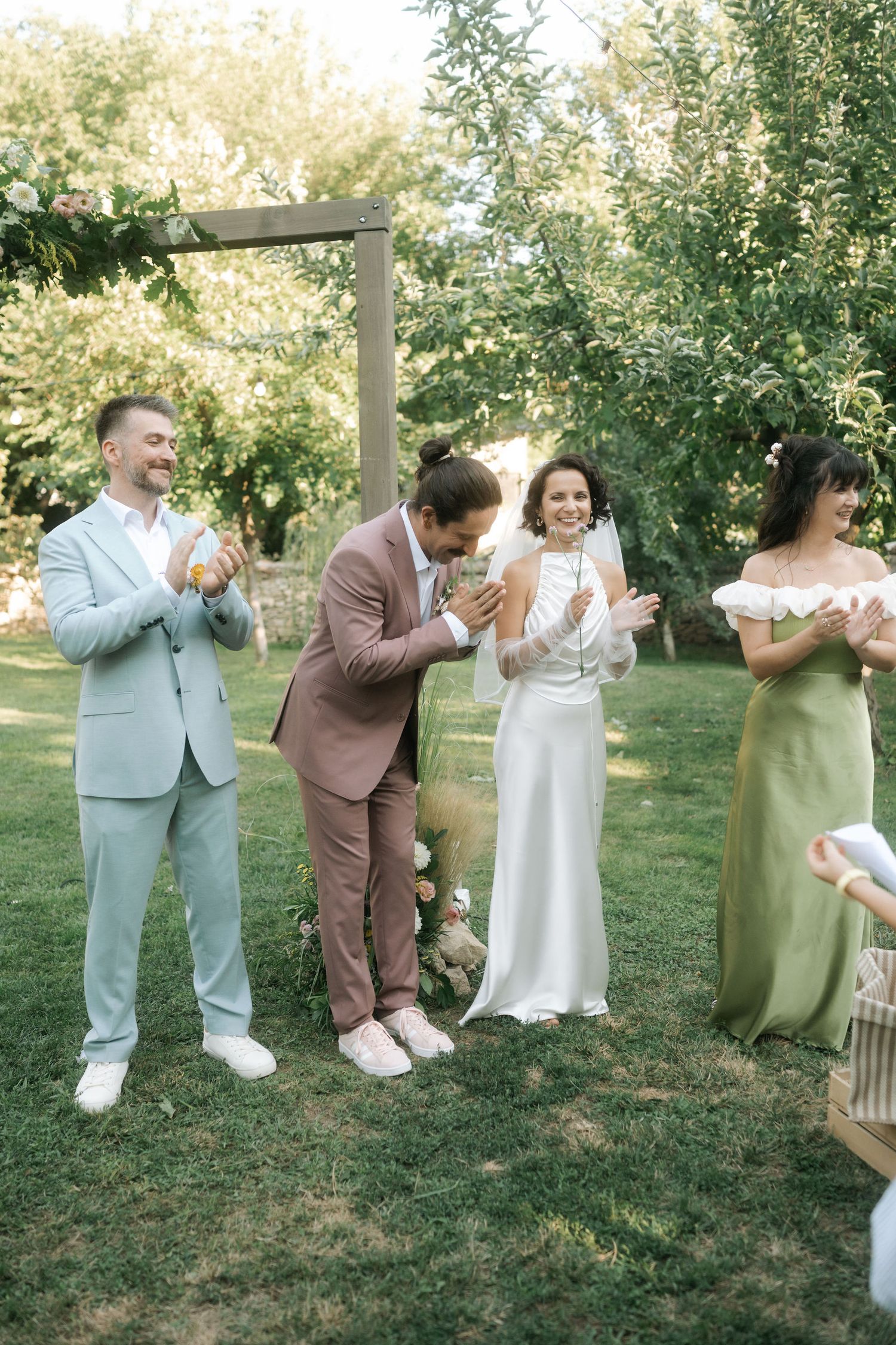 Wedding party members in pastel suits and dresses share a candid moment outdoors under trees.