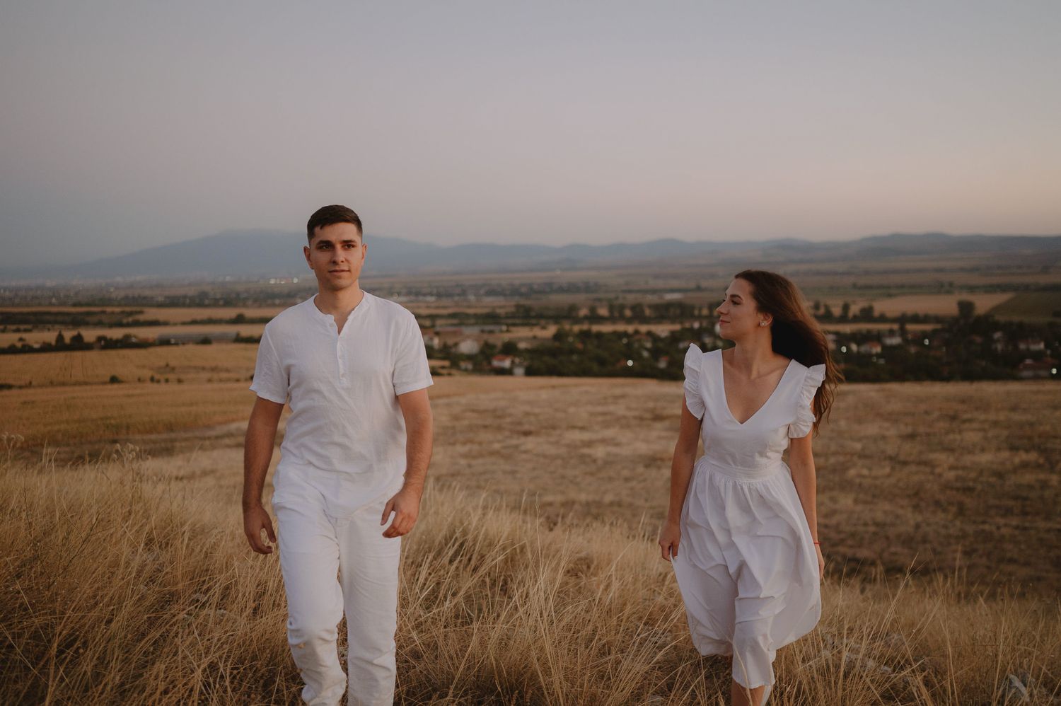 A couple in white outfits poses together in a golden wheat field during sunset with mountains in the background.
