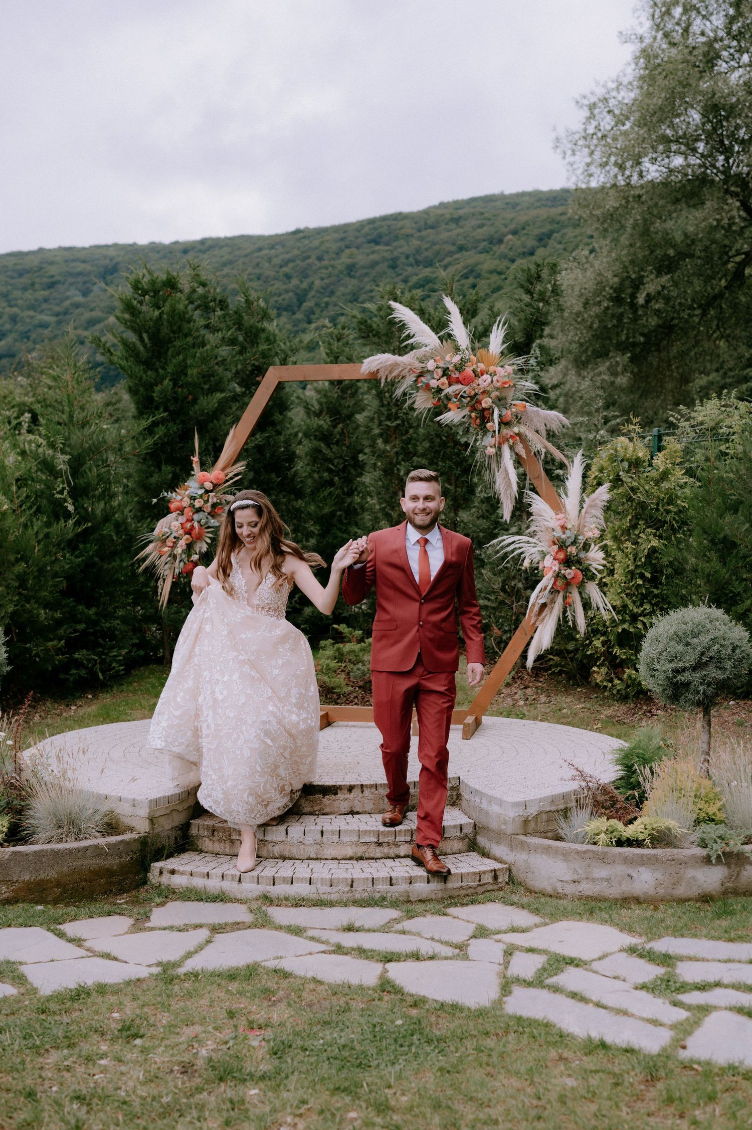 A couple poses playfully on stone steps in an outdoor wedding setting with mountains and trees in the background.