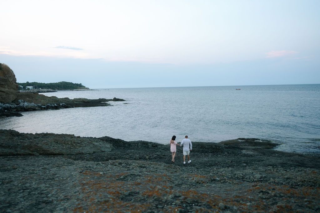 Coastal landscape panorama showing rocky shoreline and calm ocean waters at dusk with distant figures on the beach.