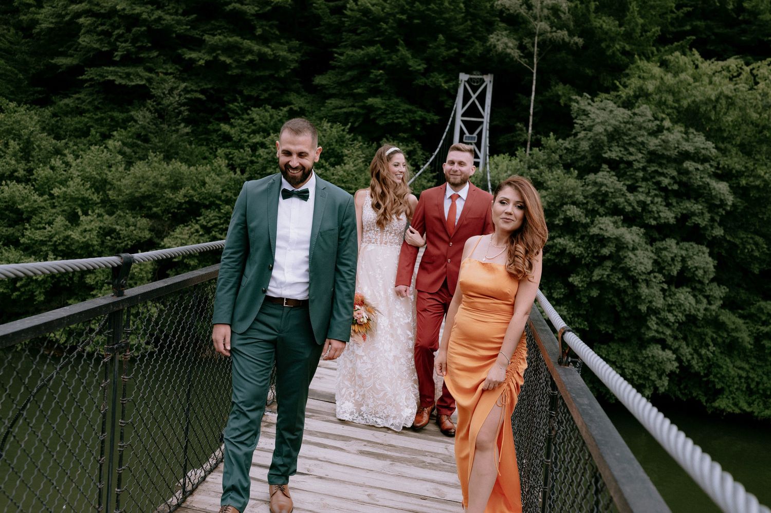 Wedding party walks across a rustic wooden suspension bridge surrounded by lush green forest.