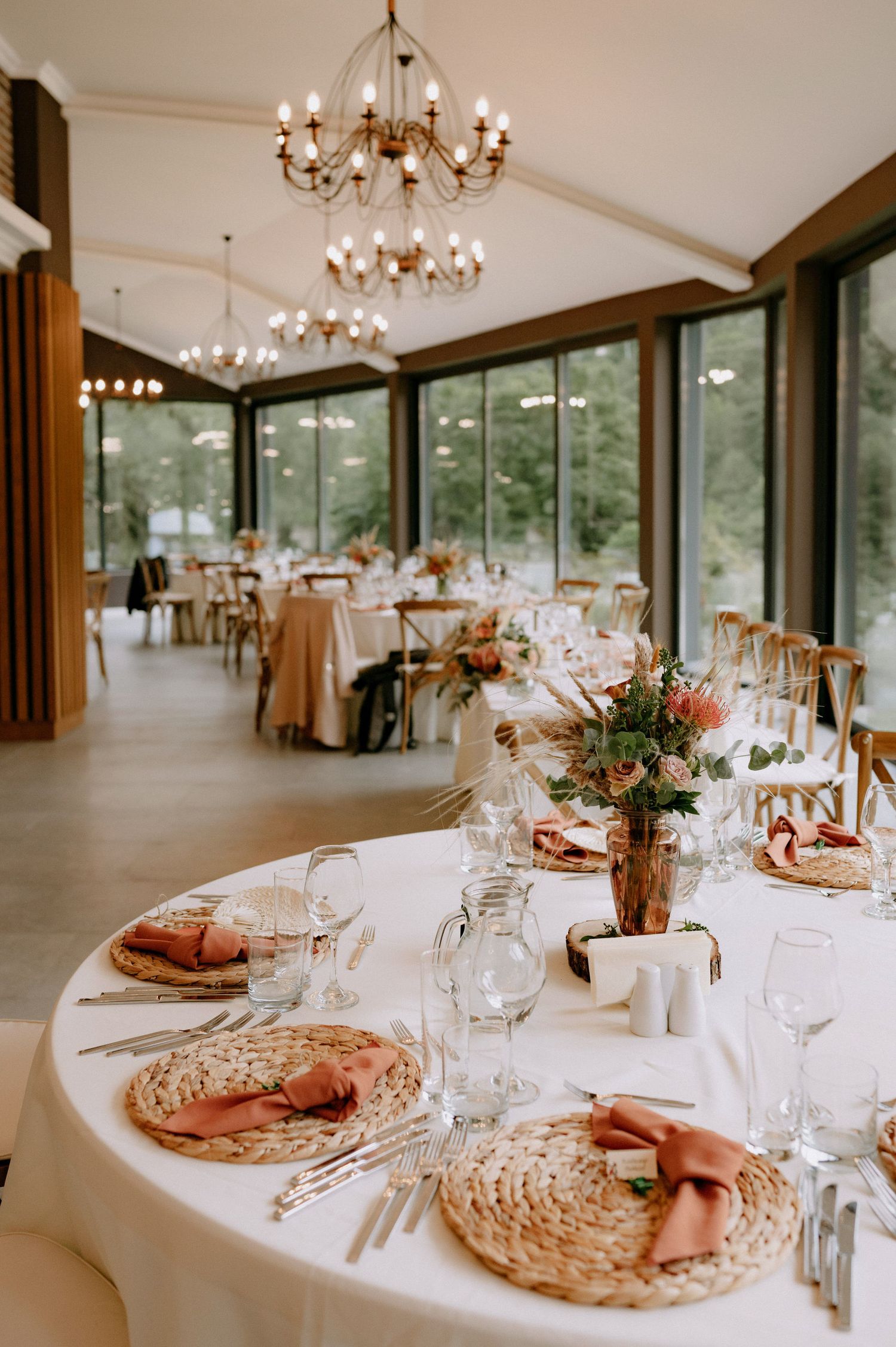 Elegant wedding reception tables set with woven charger plates, floral centerpieces and crystal chandeliers in a bright venue.