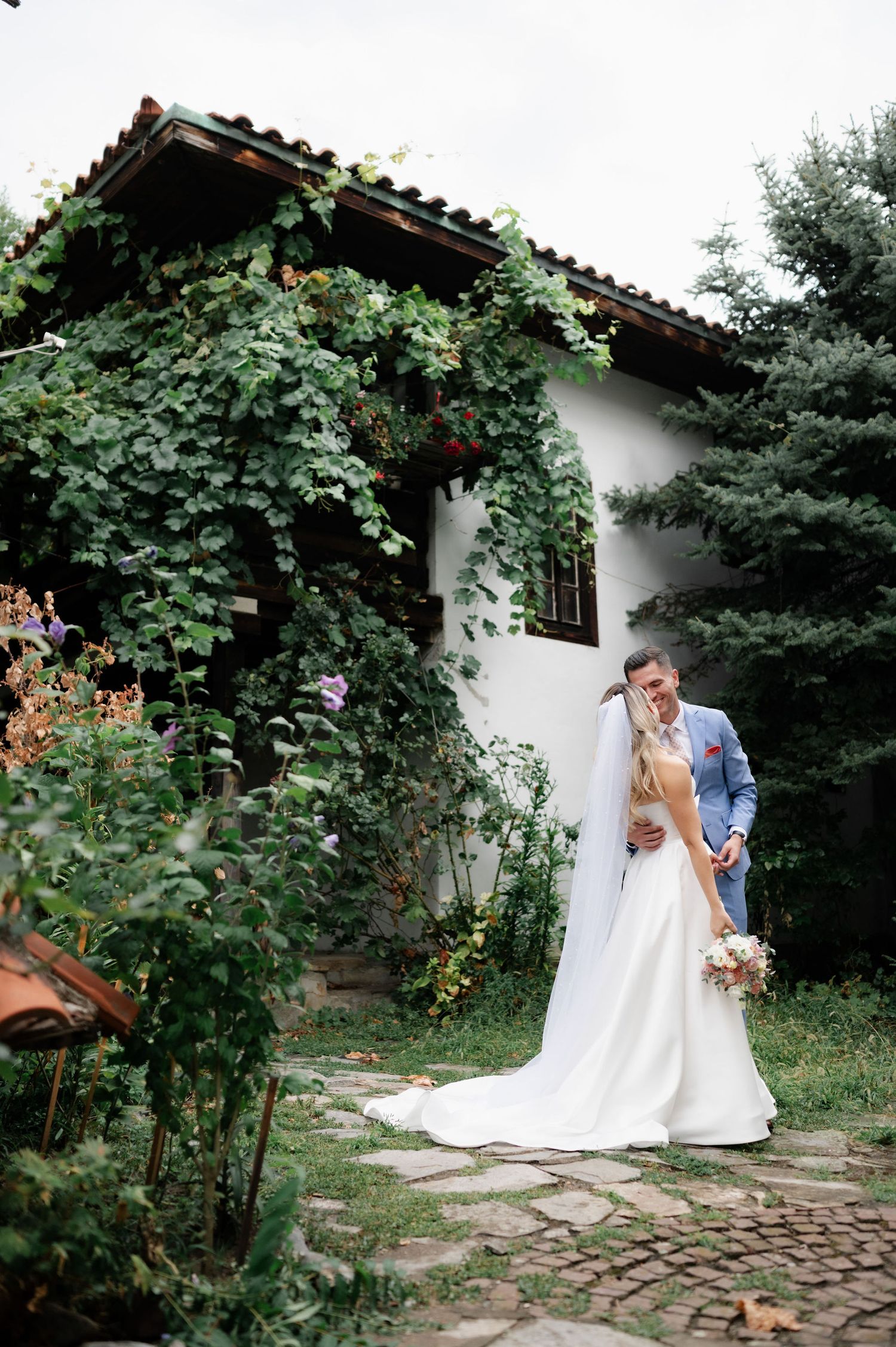 Couple in wedding attire shares romantic moments in a garden setting with stone pathway and white cottage backdrop.