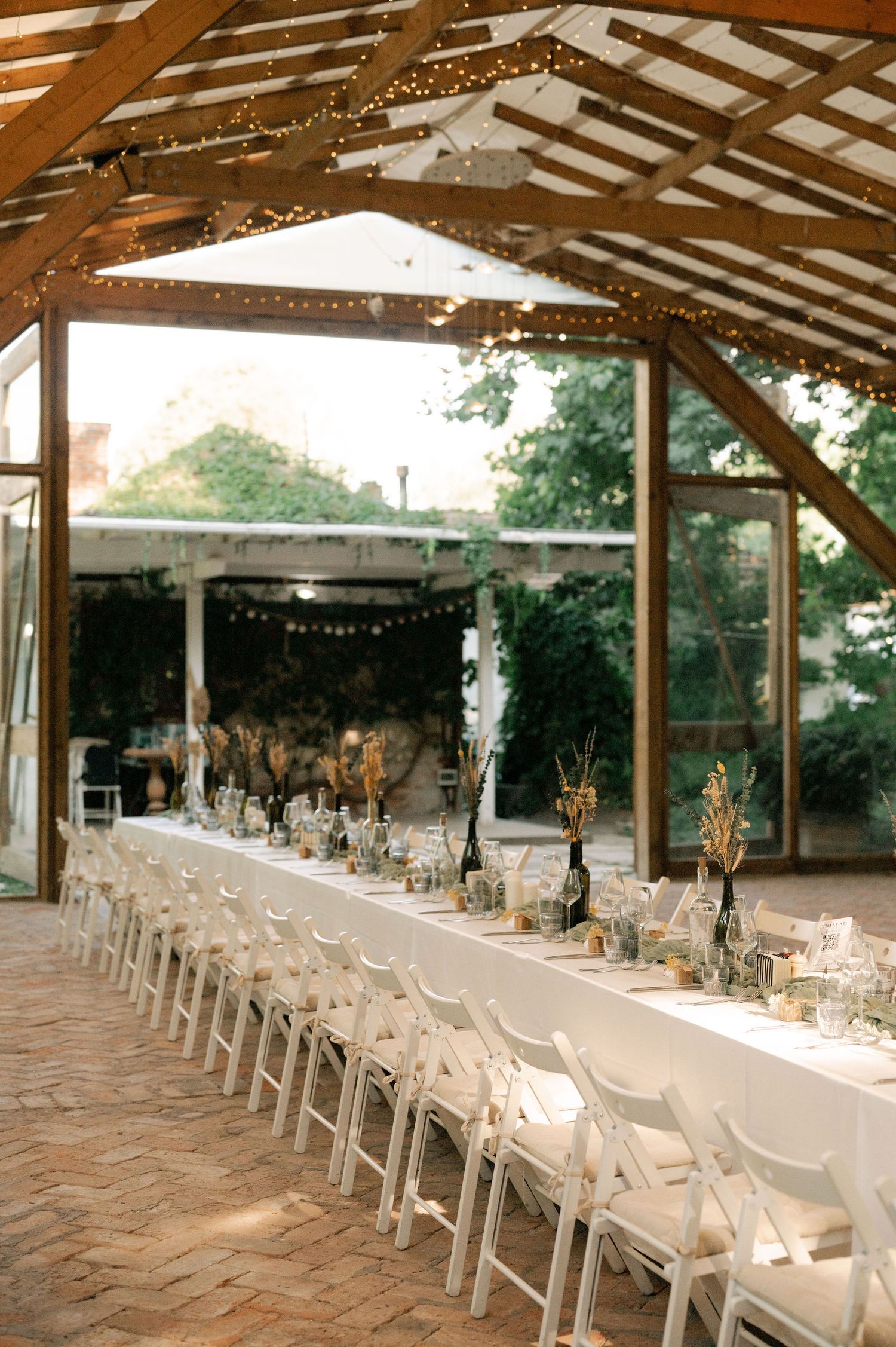 Long rustic covered patio set up for an elegant outdoor wedding reception with white chairs and string lights overhead.