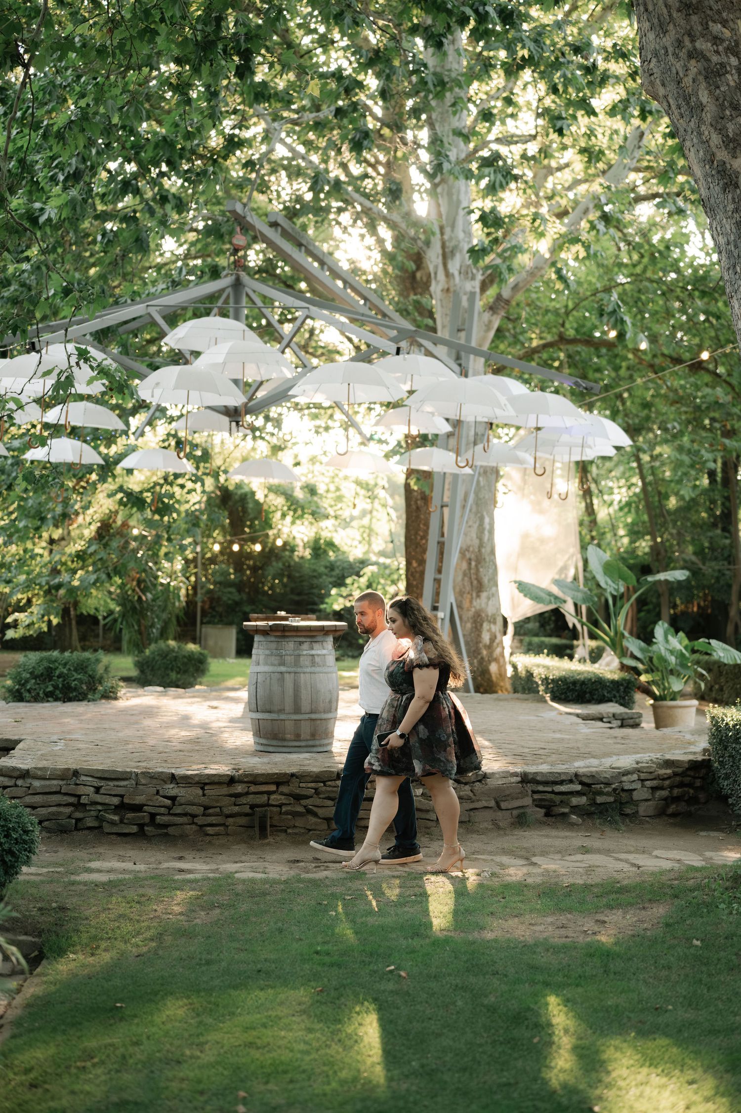 Couple embraces under white umbrellas in a lush garden setting with stone walls and green grass.