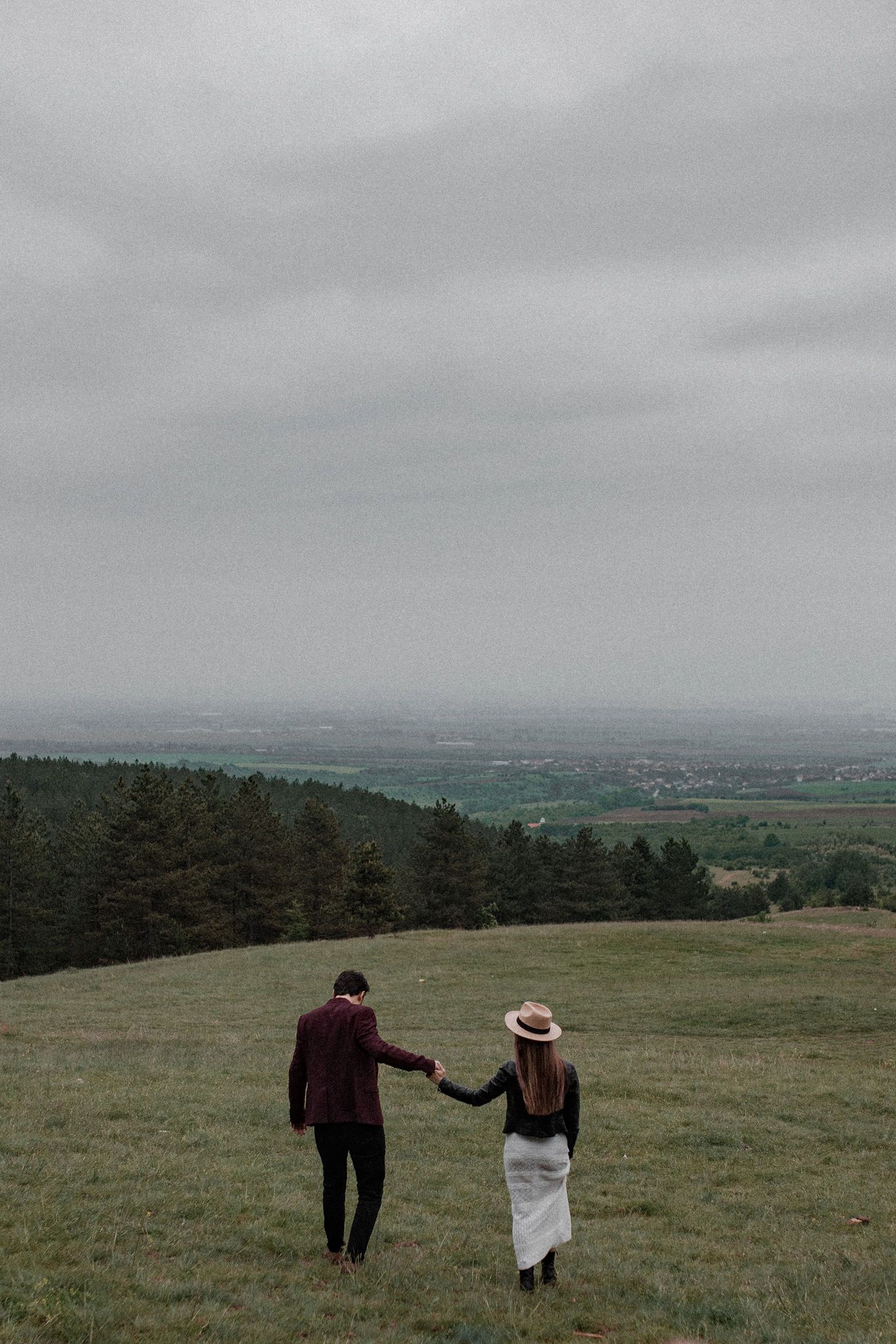 Two figures stand in a misty green meadow holding white fabric against a backdrop of distant hills and forests.