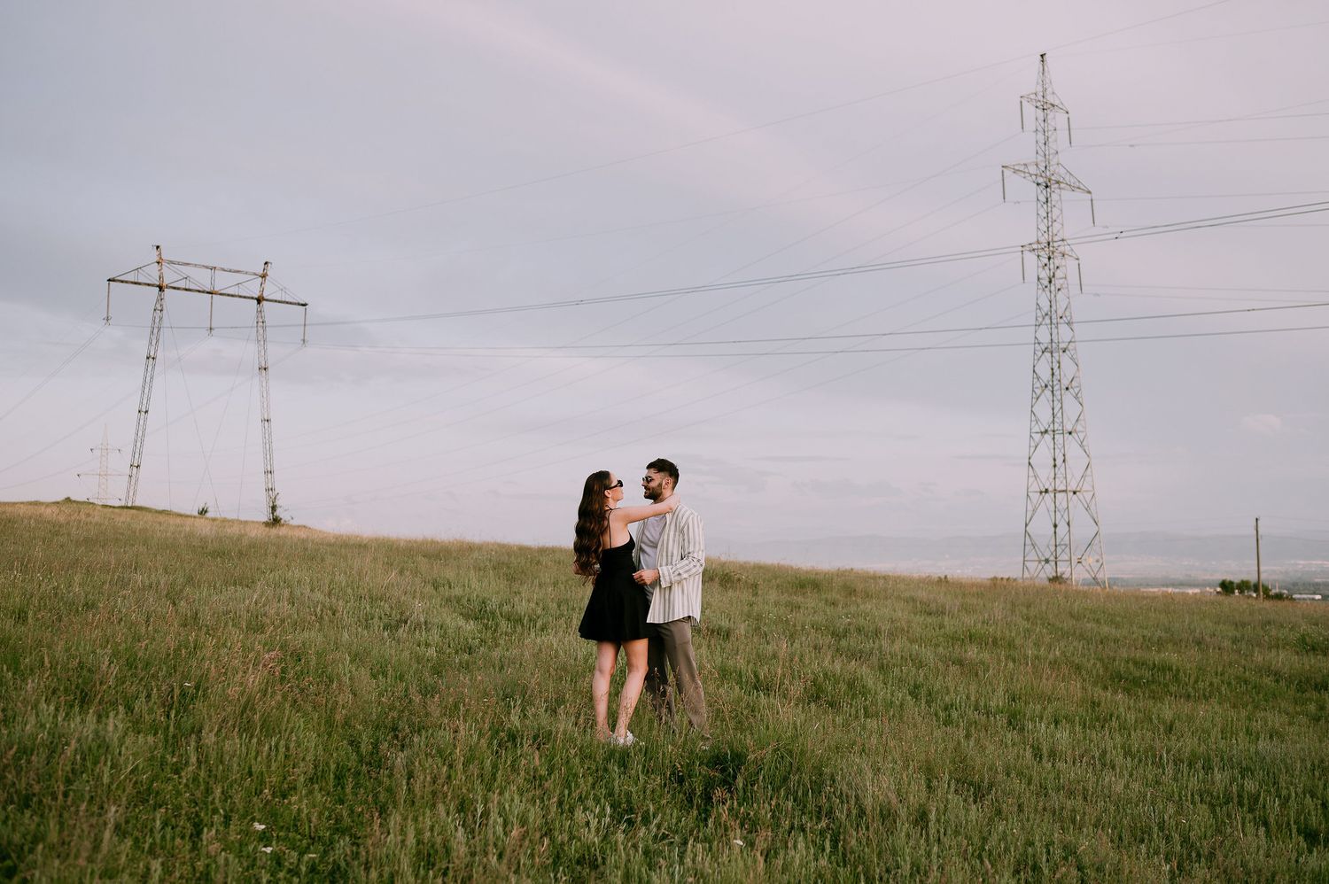 Two figures walk across a grassy hillside beneath electrical transmission towers and power lines at sunset.