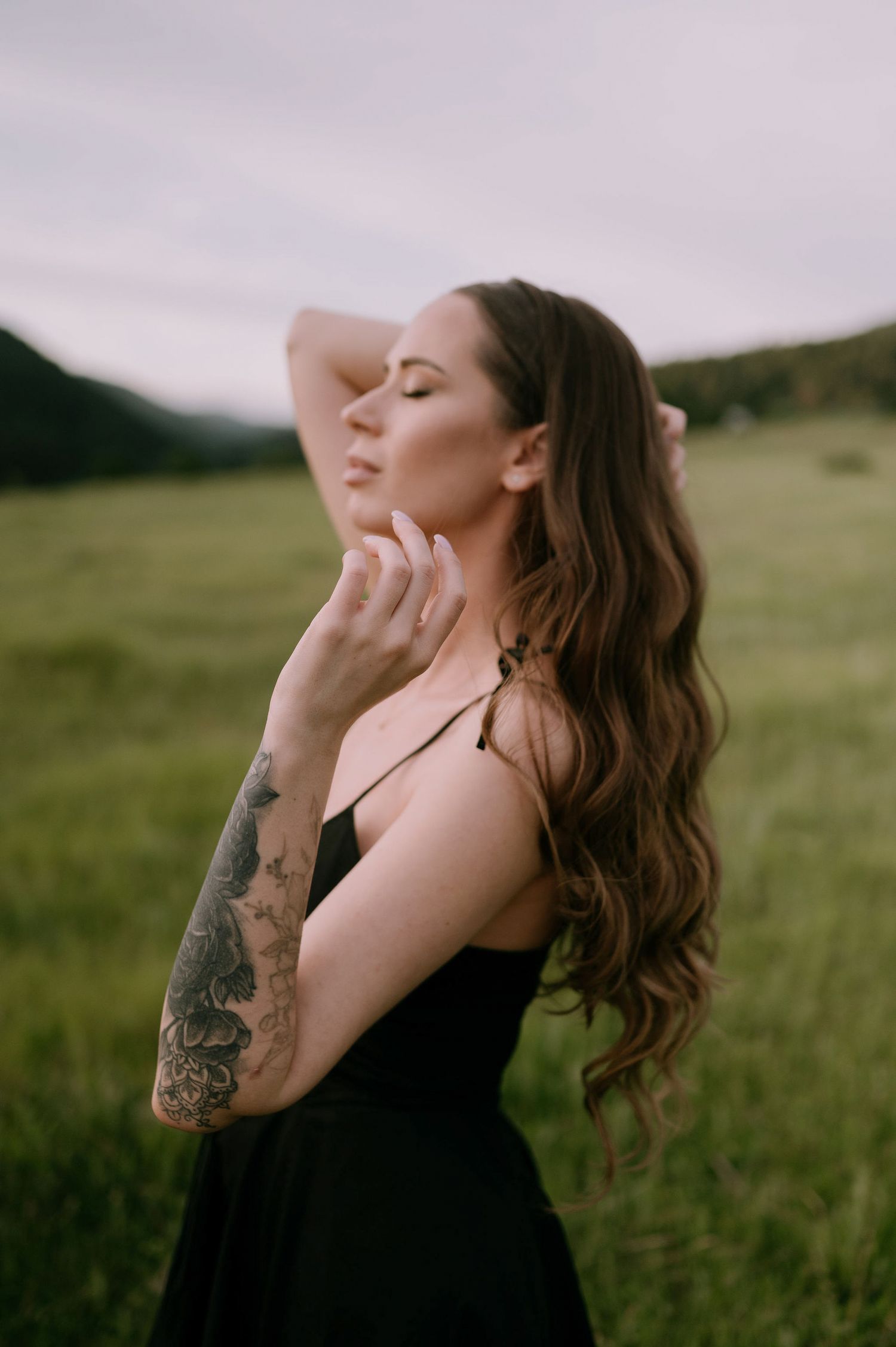 A person with long wavy hair and tattoos poses expressively in a green field wearing a black dress.
