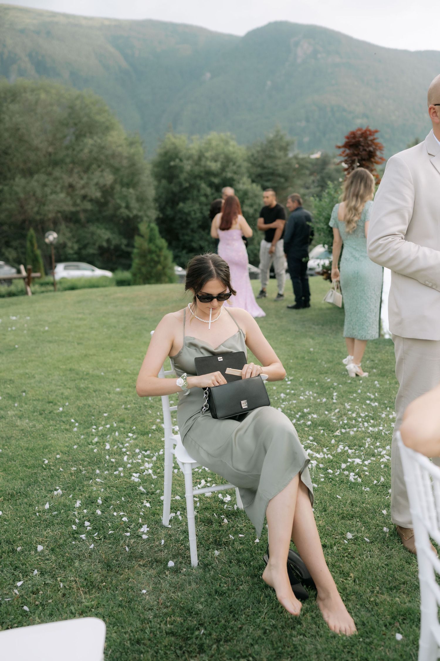 A person in a grey dress sits on a white chair in a grassy outdoor venue with mountains in the background.