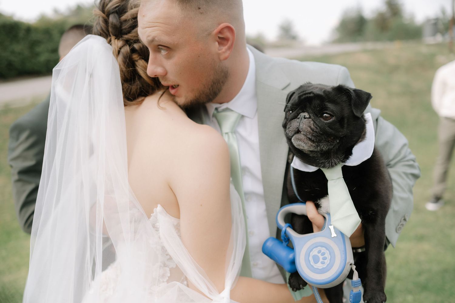 A black pug wearing a bow tie and formal attire stands with a couple during their wedding ceremony outdoors.