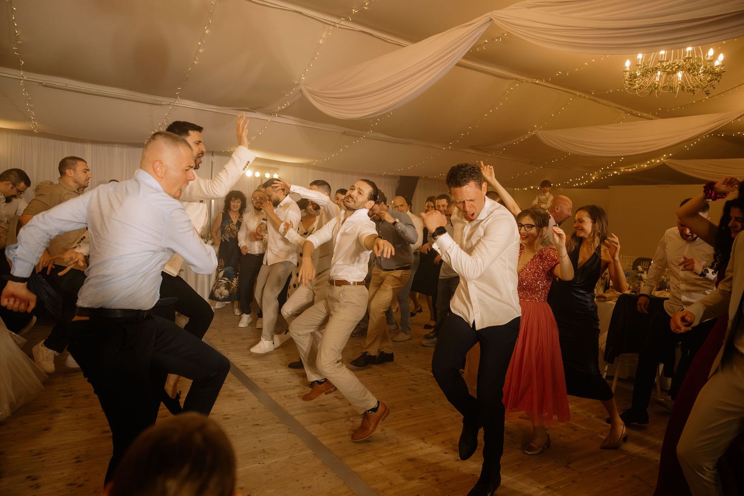 Guests dancing energetically at a wedding reception in an elegant ballroom with draped ceiling decor.