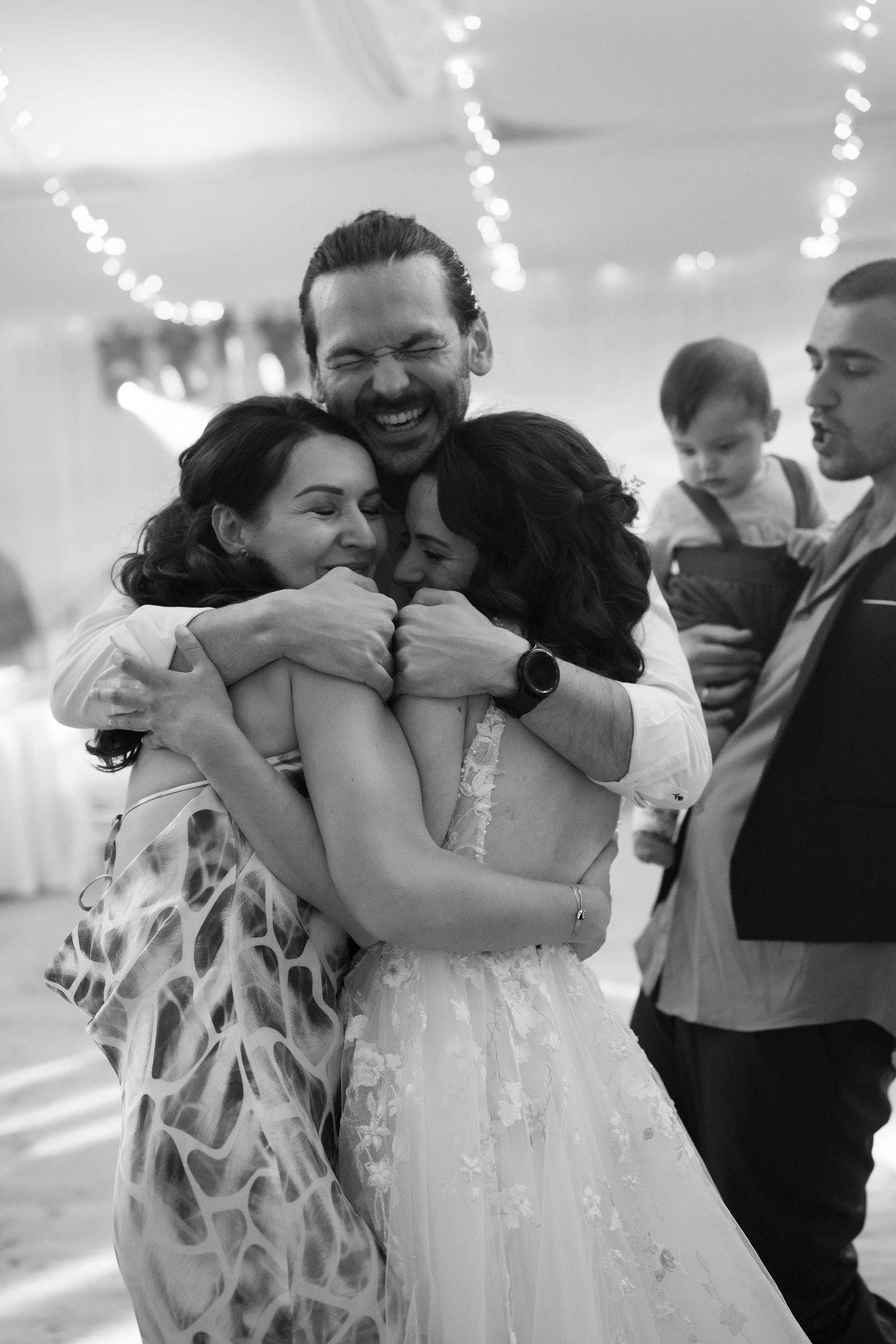 Black and white photo of a joyful group hug at a wedding reception, with a smiling man embracing two women in elegant attire.