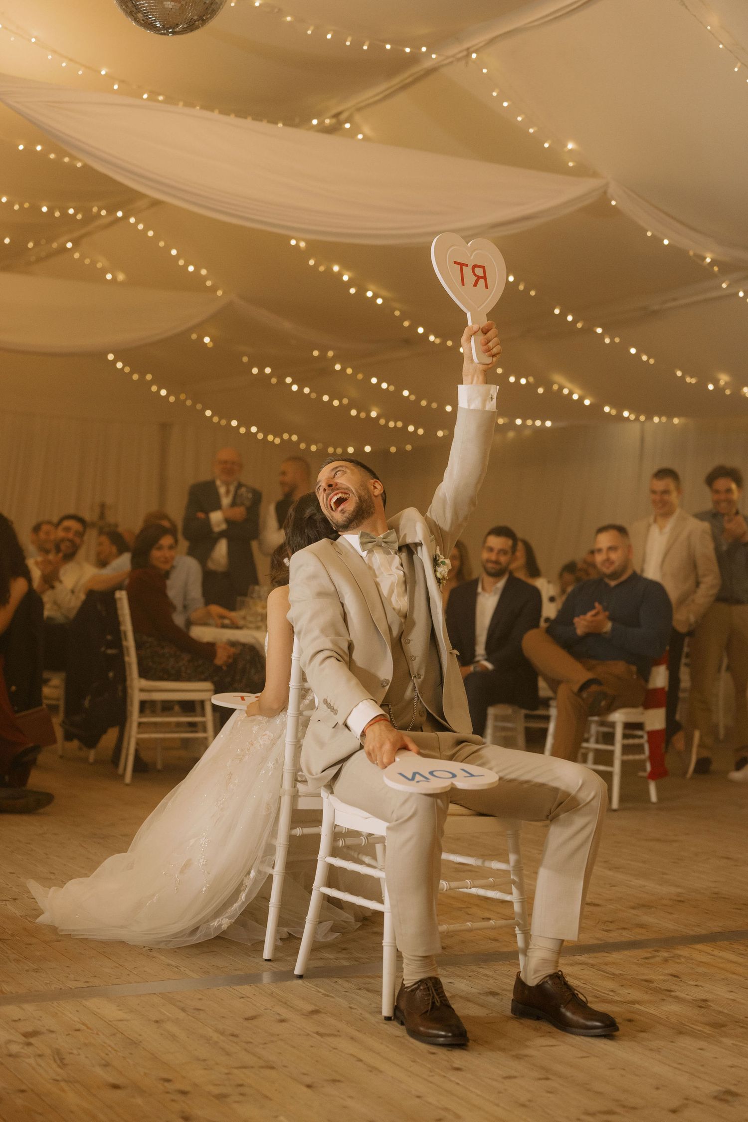 Groom in gray suit raises heart sign while bride laughs beside him during fun wedding game at decorated reception venue.