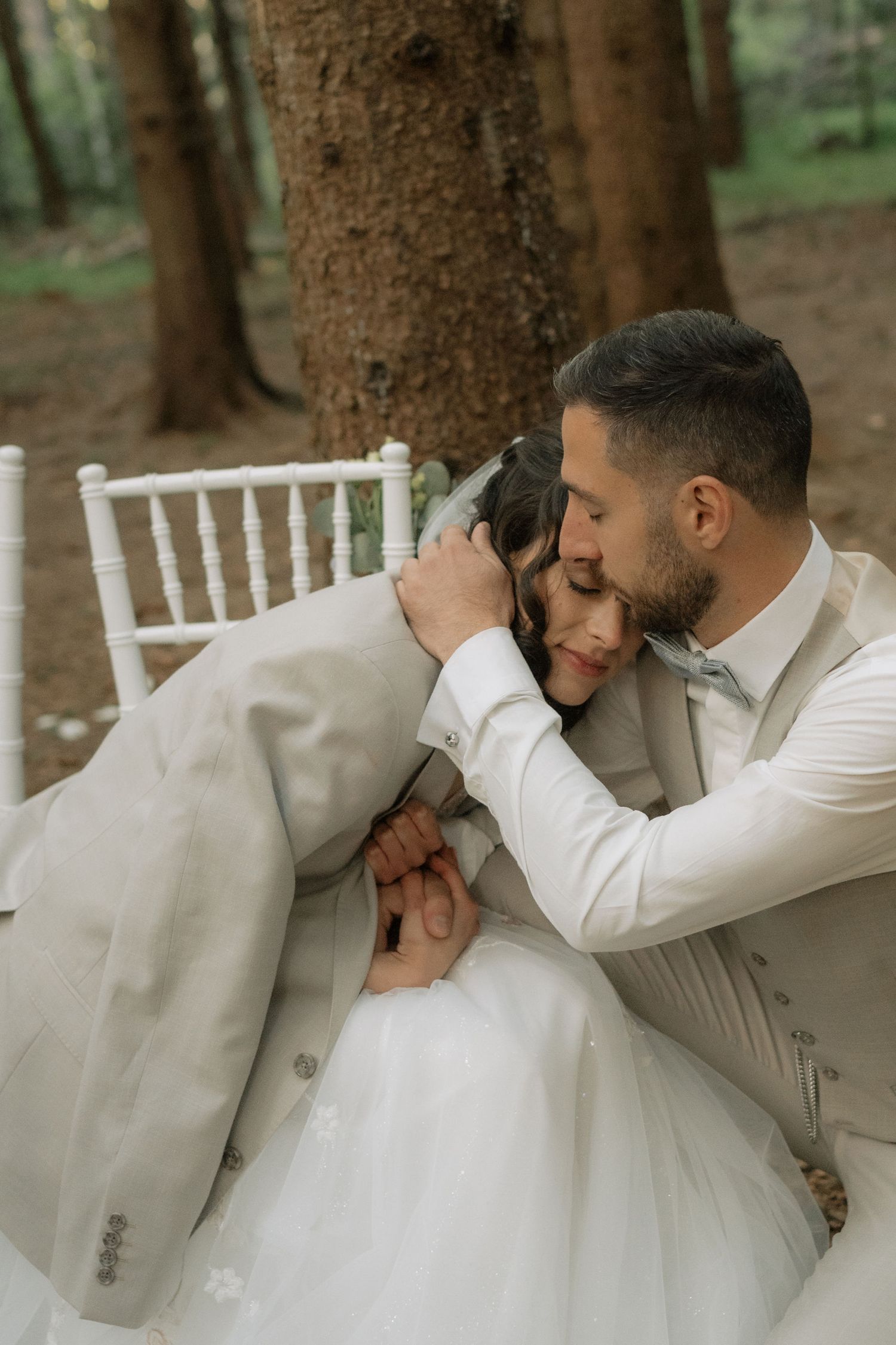 Groom in white suit embraces bride in wedding dress among forest trees, sharing a romantic intimate moment.